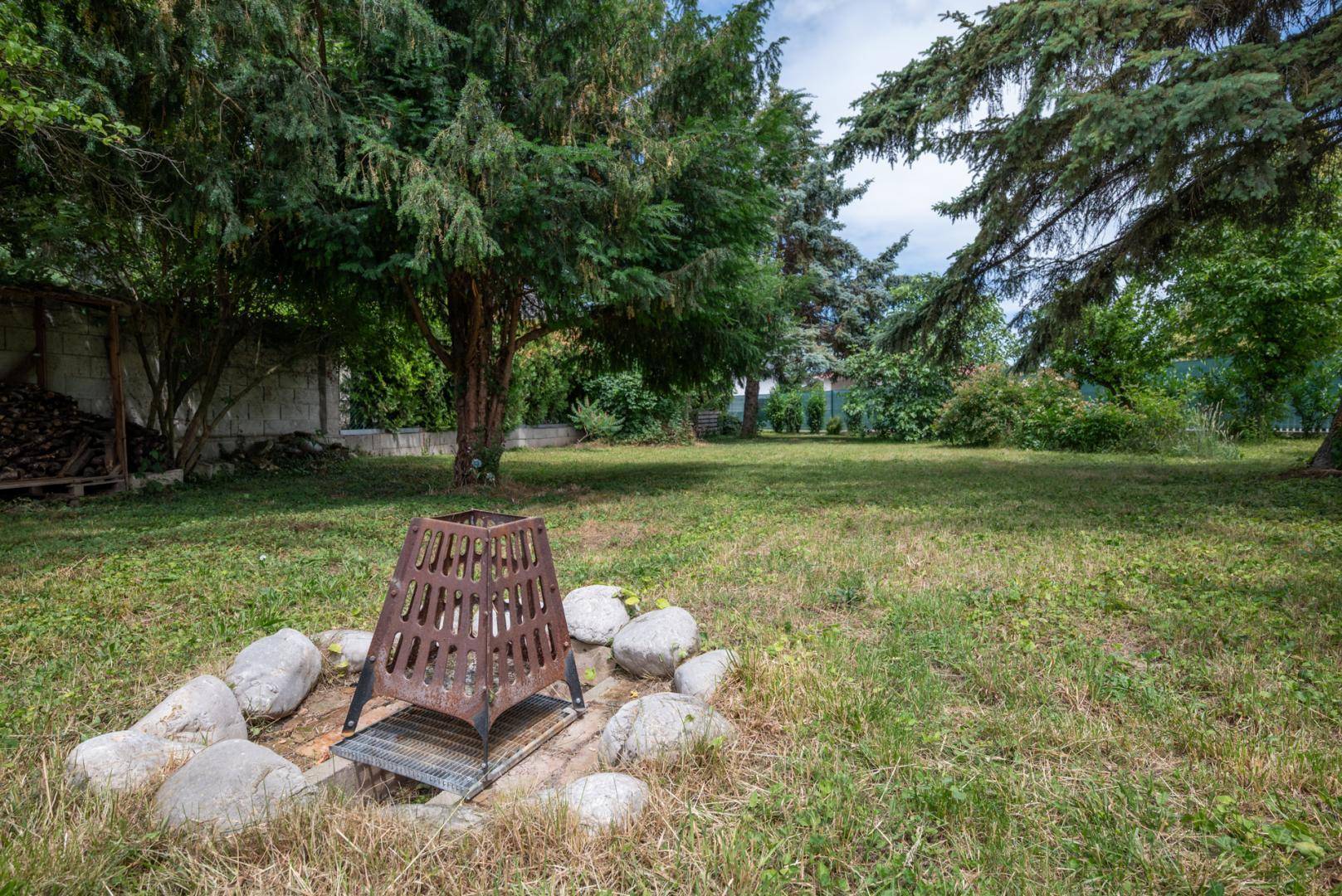 Großer Gartenbereich mit einer Feuerstelle umgeben von Steinen und altem Baumbestand.