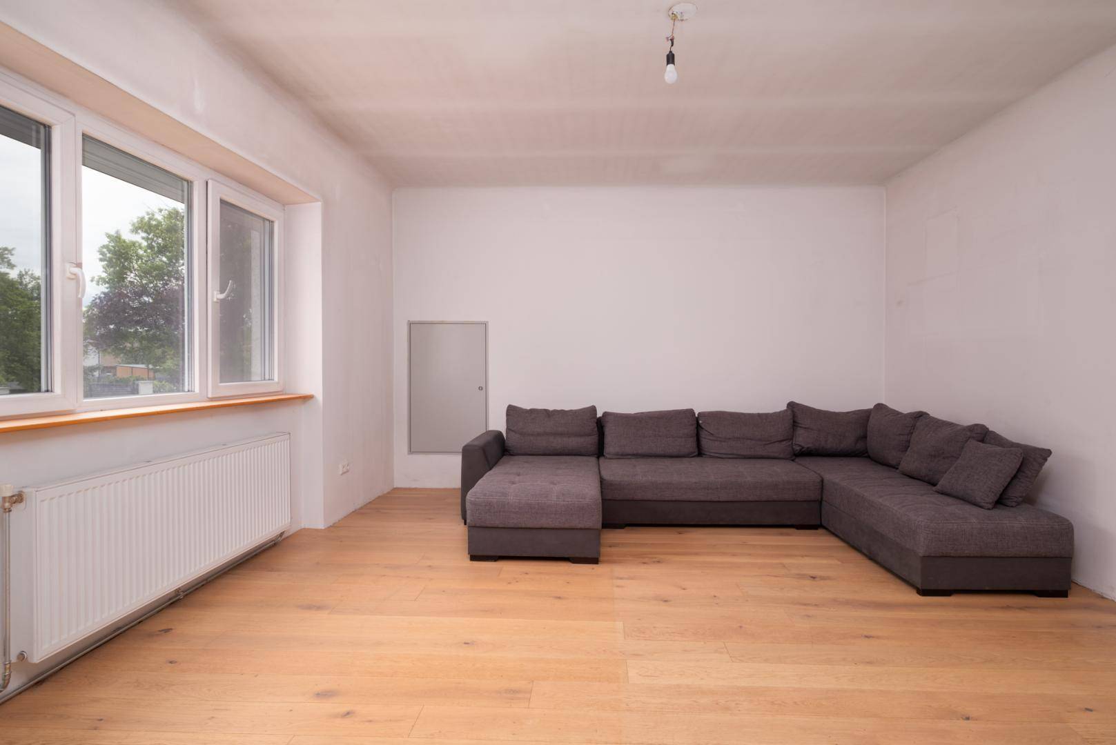 Geräumiges Wohnzimmer mit hellem Holzboden, großem Fenster und einer modernen grauen Eckcouch.