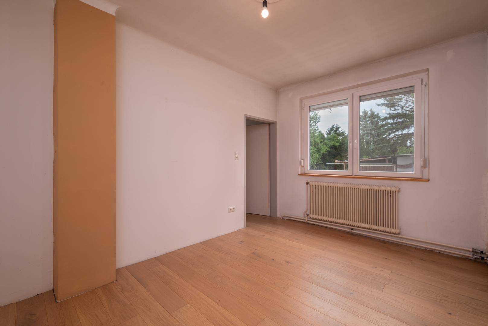 Leeres Schlafzimmer mit hellem Holzboden, großem Fenster und Heizkörper, bereit zur Einrichtung.