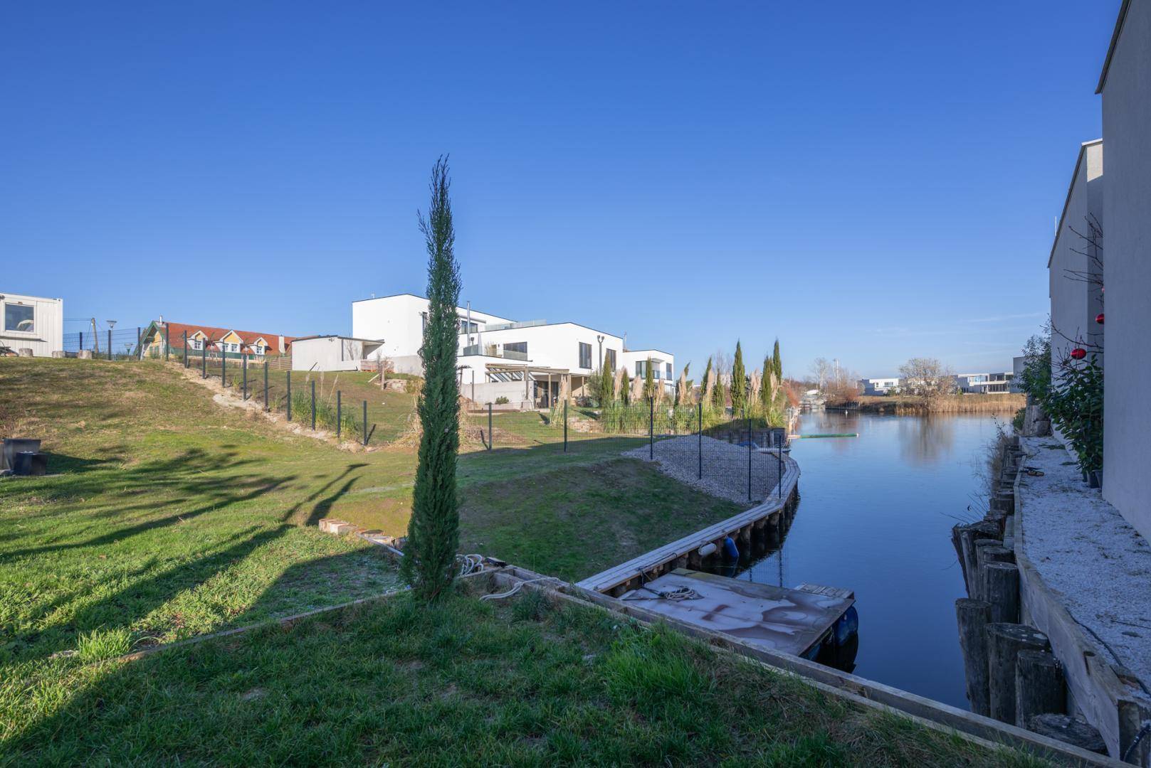 Kanalufer mit Anlegesteg, umgeben von grünen Hängen und modernen Wohngebäuden unter blauem Himmel.