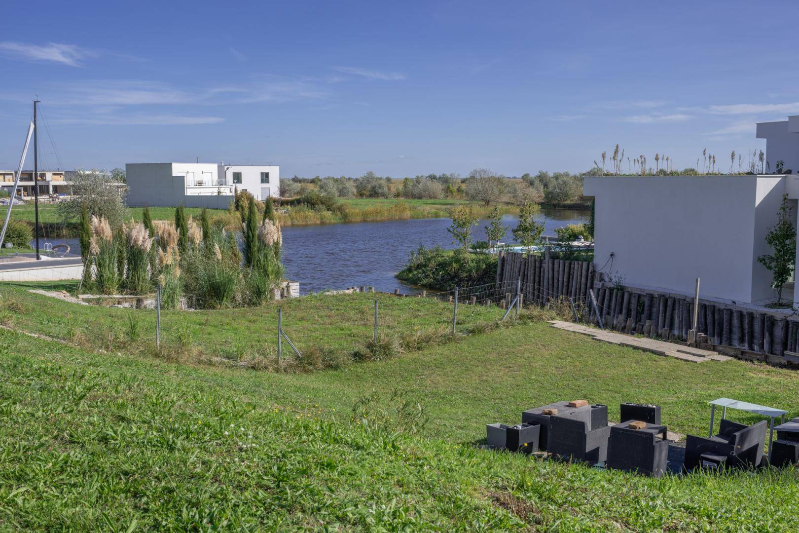 Grüne Außenfläche mit einfachen Sitzgelegenheiten und Blick auf den Kanal sowie moderne Gebäude.
