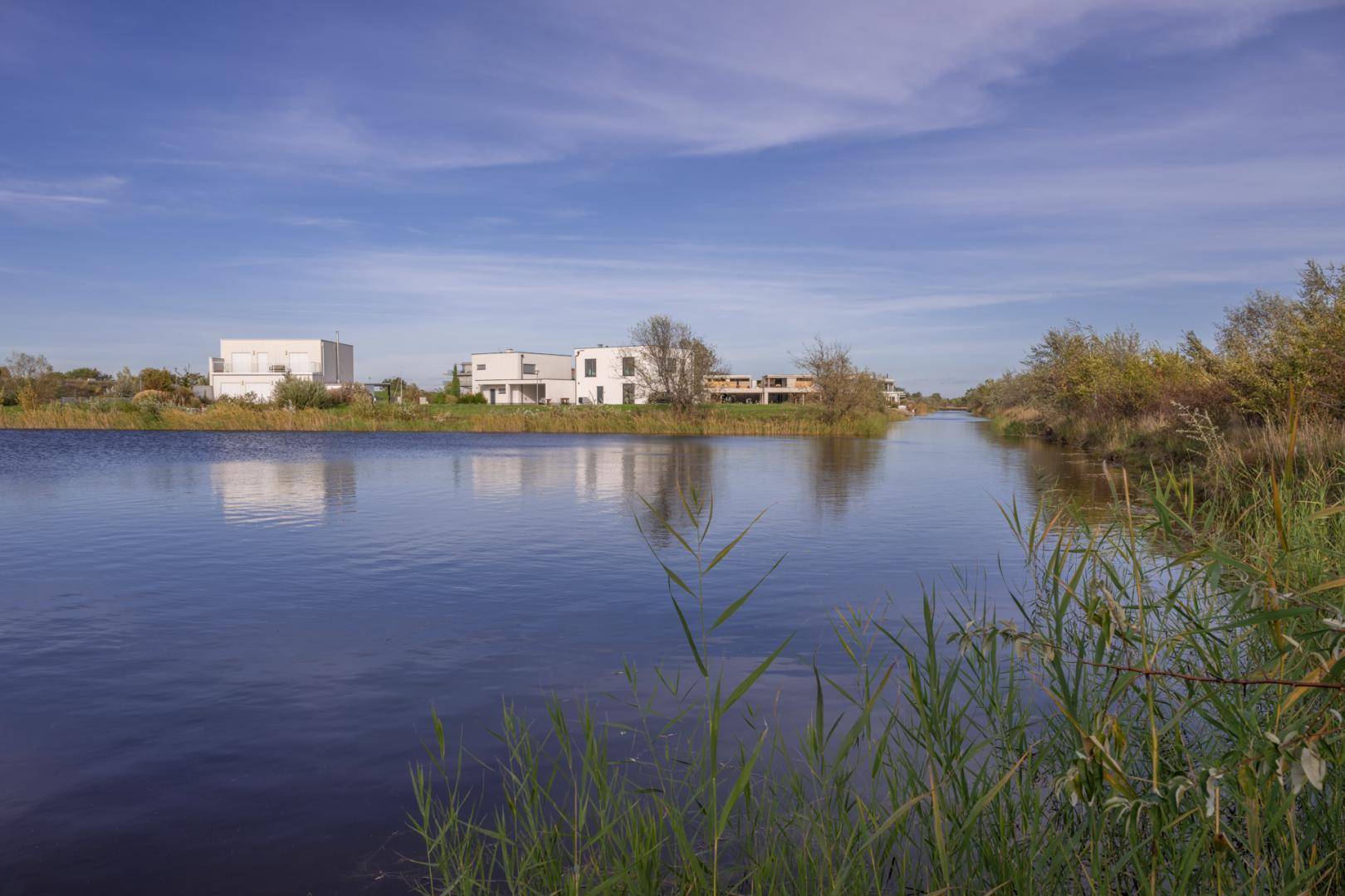 Ruhiger Kanal mit modernen Häusern am Ufer, umgeben von Schilf und weitem Grün der Landschaft.