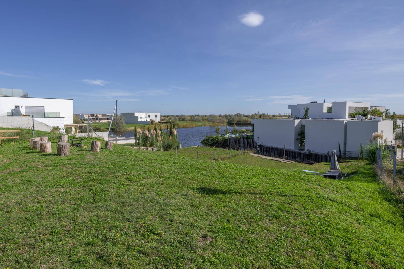 Weitläufige Grünfläche mit rustikalen Sitzgelegenheiten und Blick auf das Wasser und moderne Häuser.