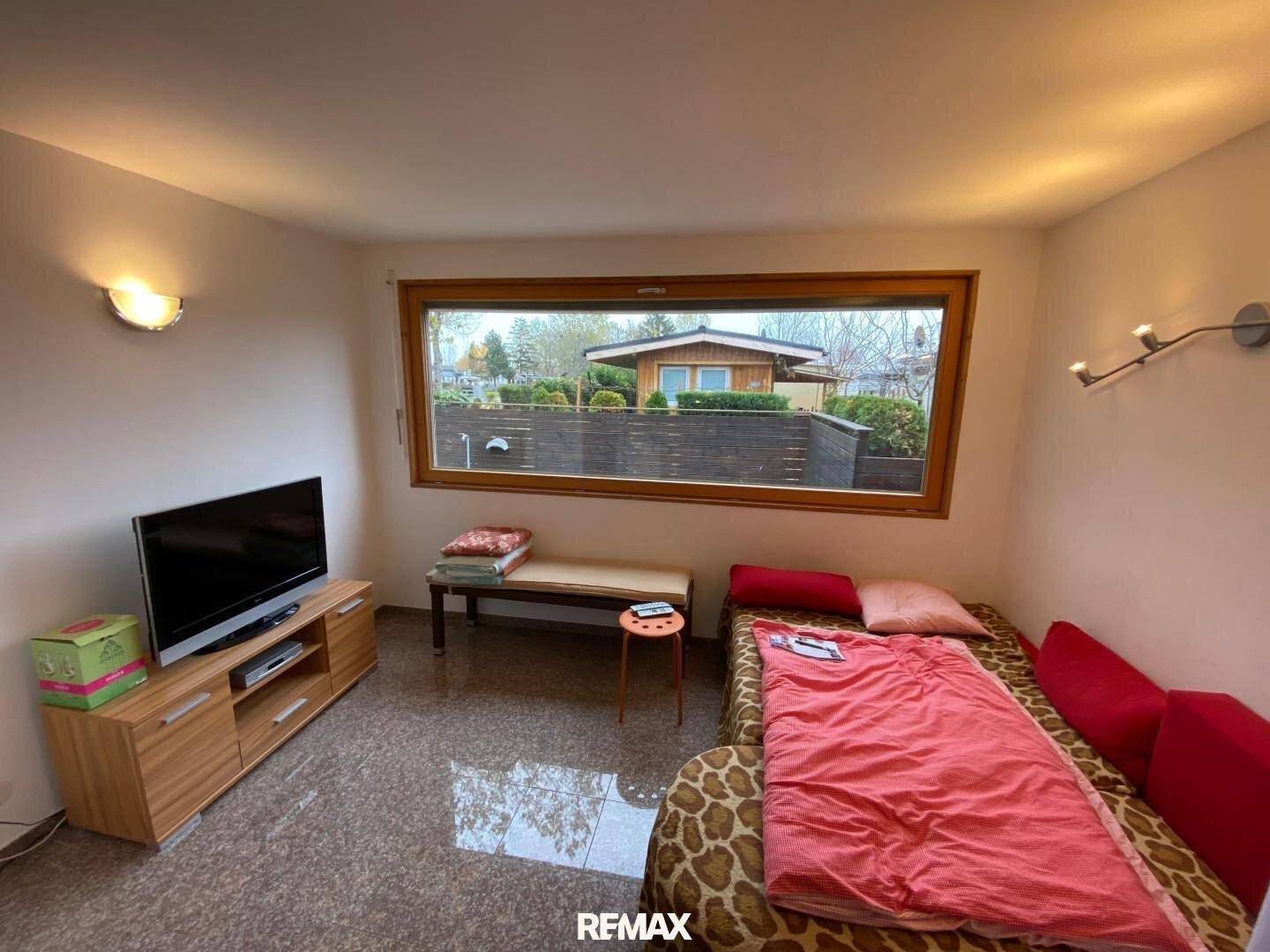 Wohnbereich mit Einzelbett, Fernseher und einem Fenster mit Blick ins Grüne.
