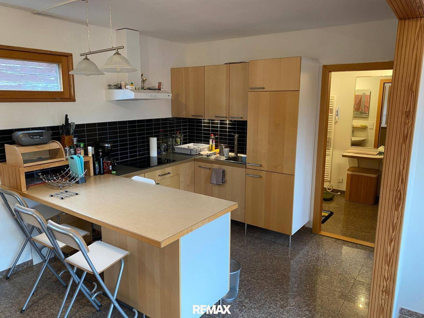 Küche mit Holzschränken, schwarzem Fliesenspiegel und Blick in ein angrenzendes Badezimmer.