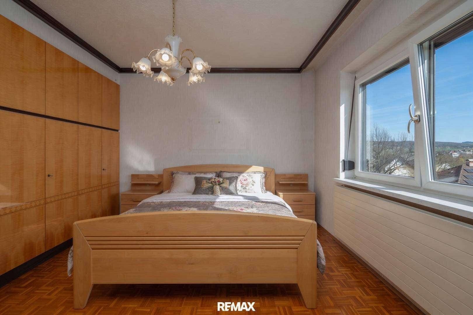 Helles Schlafzimmer mit Holzmöbeln, Parkettboden und Fenster mit Blick ins Grüne.
