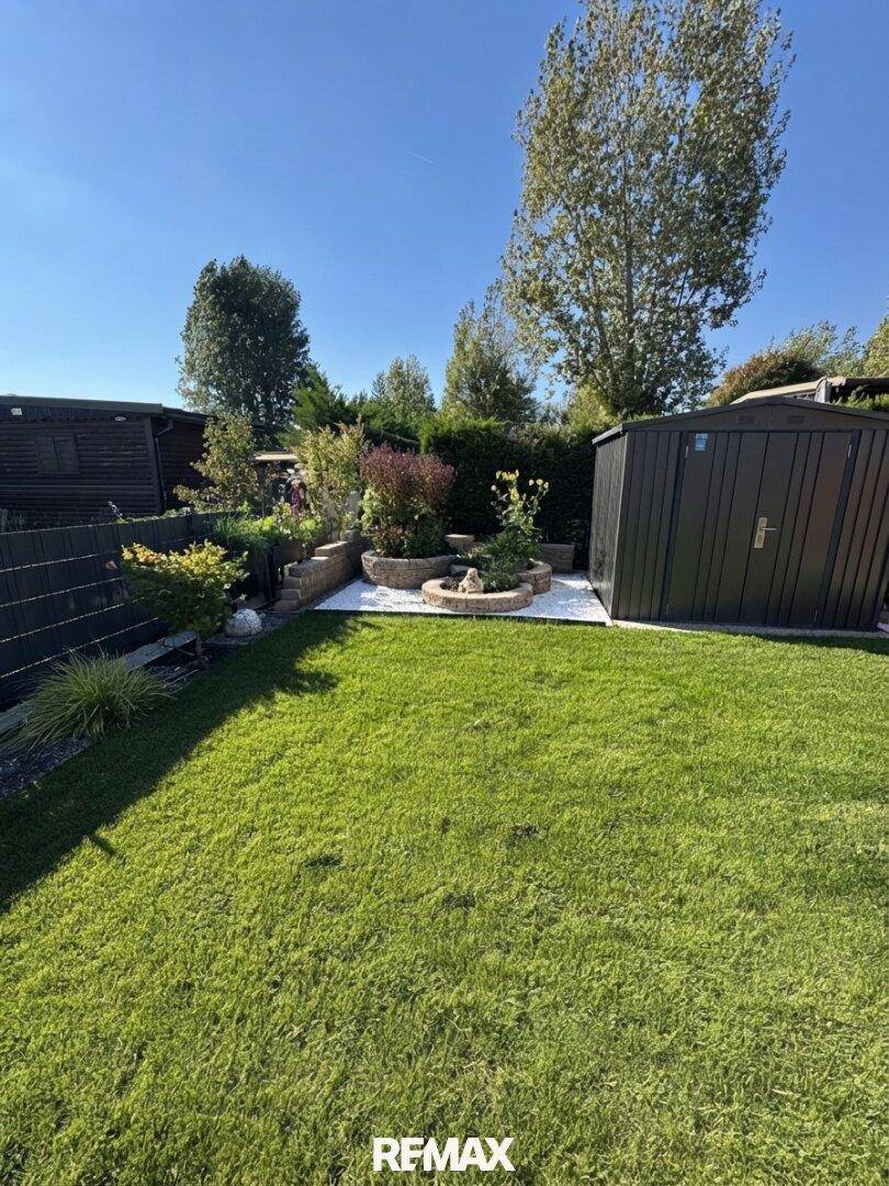 Gepflegter Gartenbereich mit Rasen, einem Gartenhaus und dekorativen Pflanzenbeeten unter blauem Himmel.
