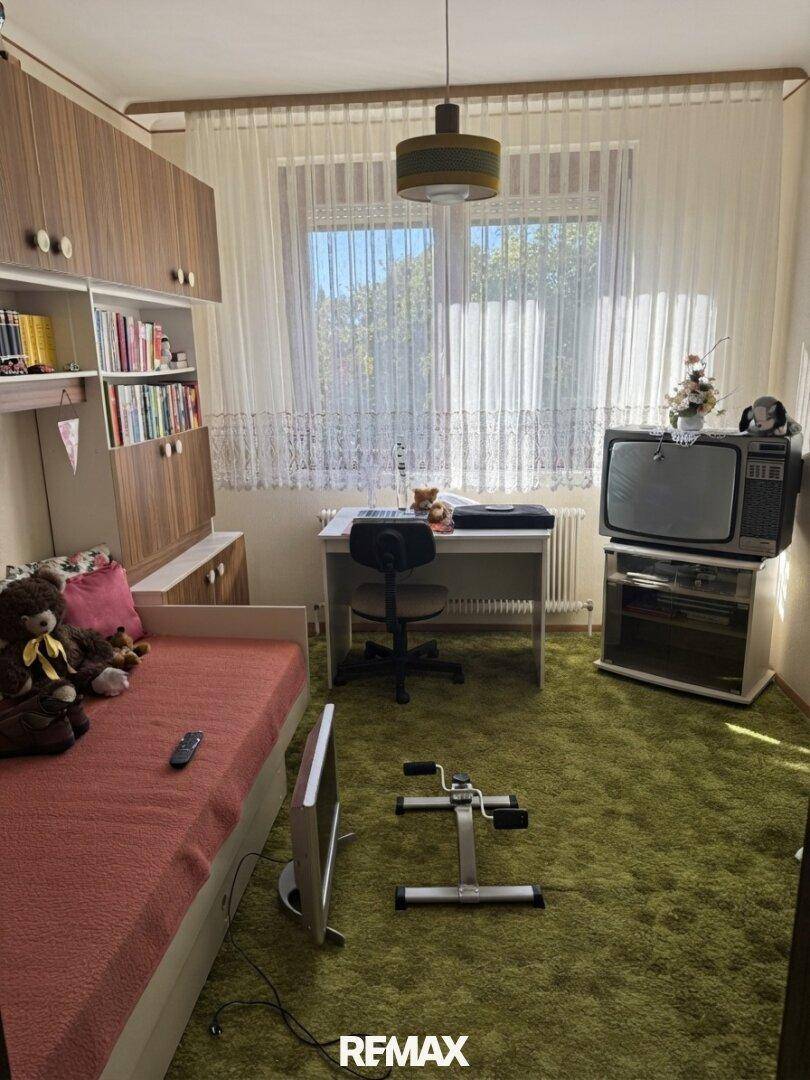 Schlafzimmer mit grünem Teppichboden, einem Bett, Schreibtisch und älteren Möbeln.