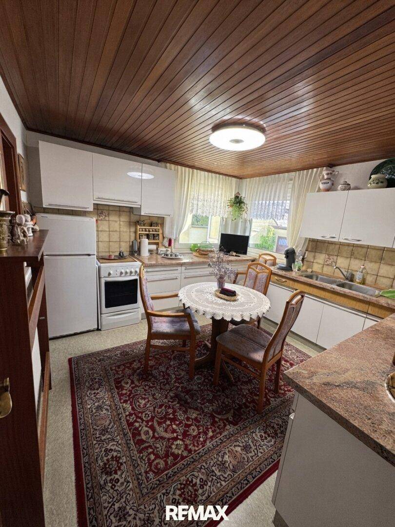 Gemütliche Küche mit Holztäfelung an der Decke, weißer Einbauküche und einem kleinen Esstisch in der Mitte.
