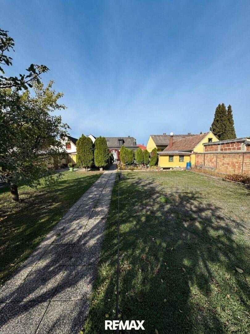 Großer, gepflegter Garten mit Rasenfläche, gepflastertem Weg und hohen Bäumen unter blauem Himmel.