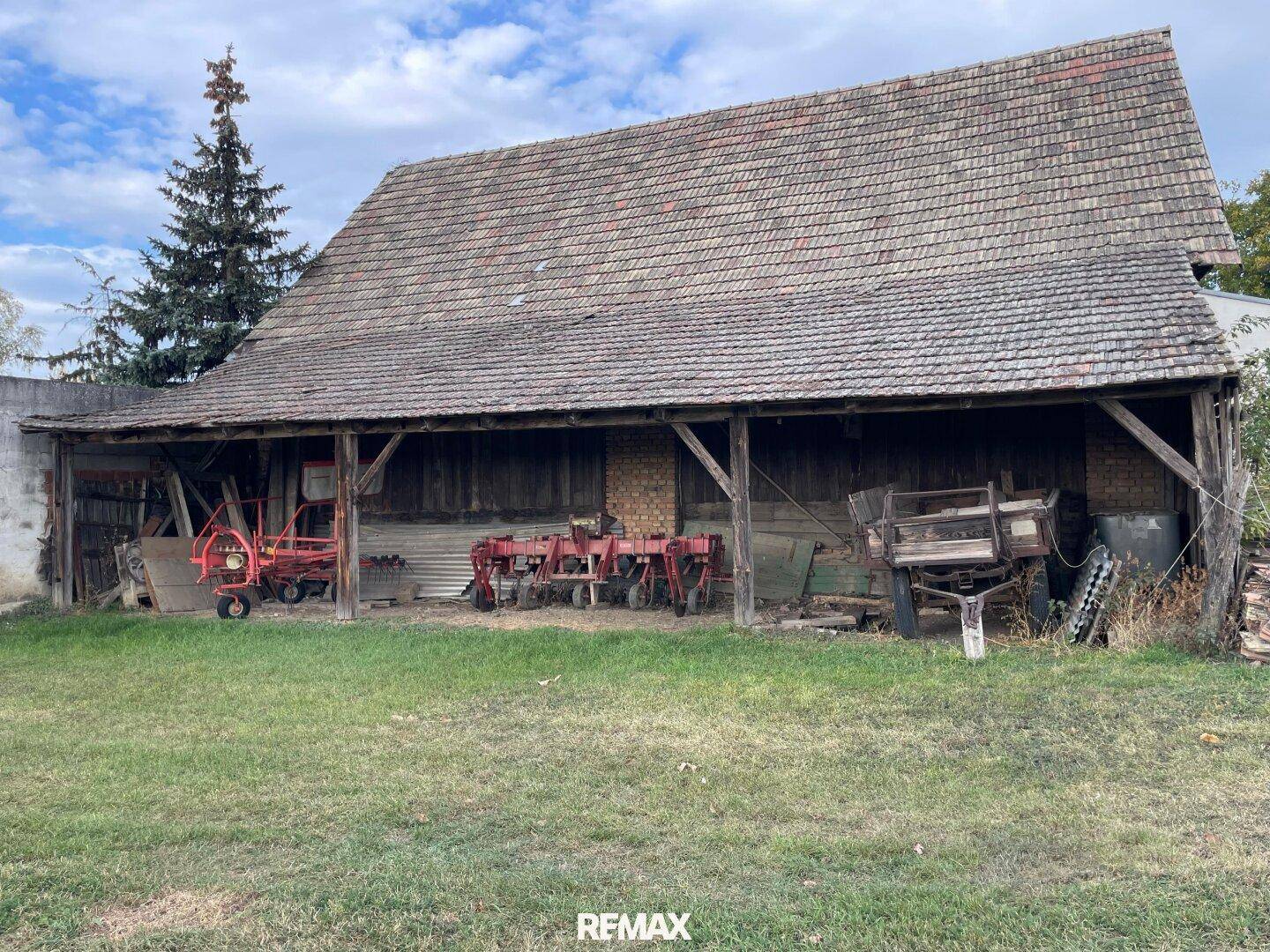 Alte offene Scheune mit Holzbalken und landwirtschaftlichen Geräten auf einem grünen Feld unter blauem Himmel.