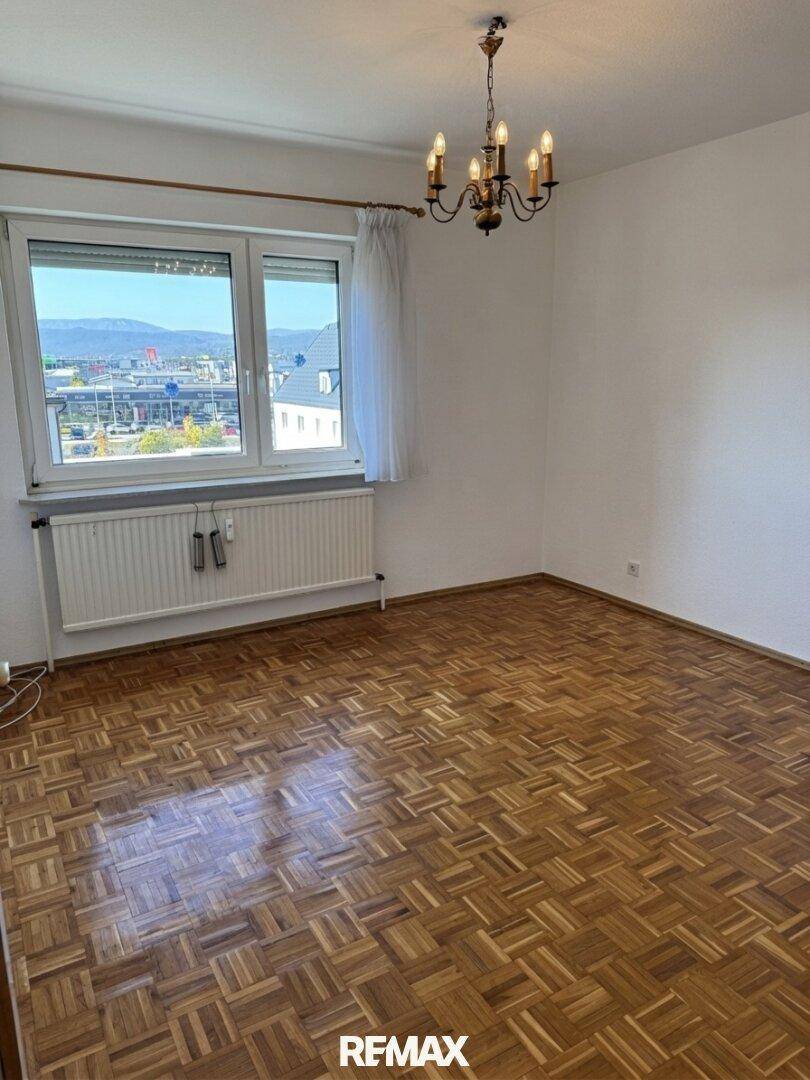 Helles Zimmer mit Parkettboden, großem Fenster und einem klassischen Deckenleuchter.