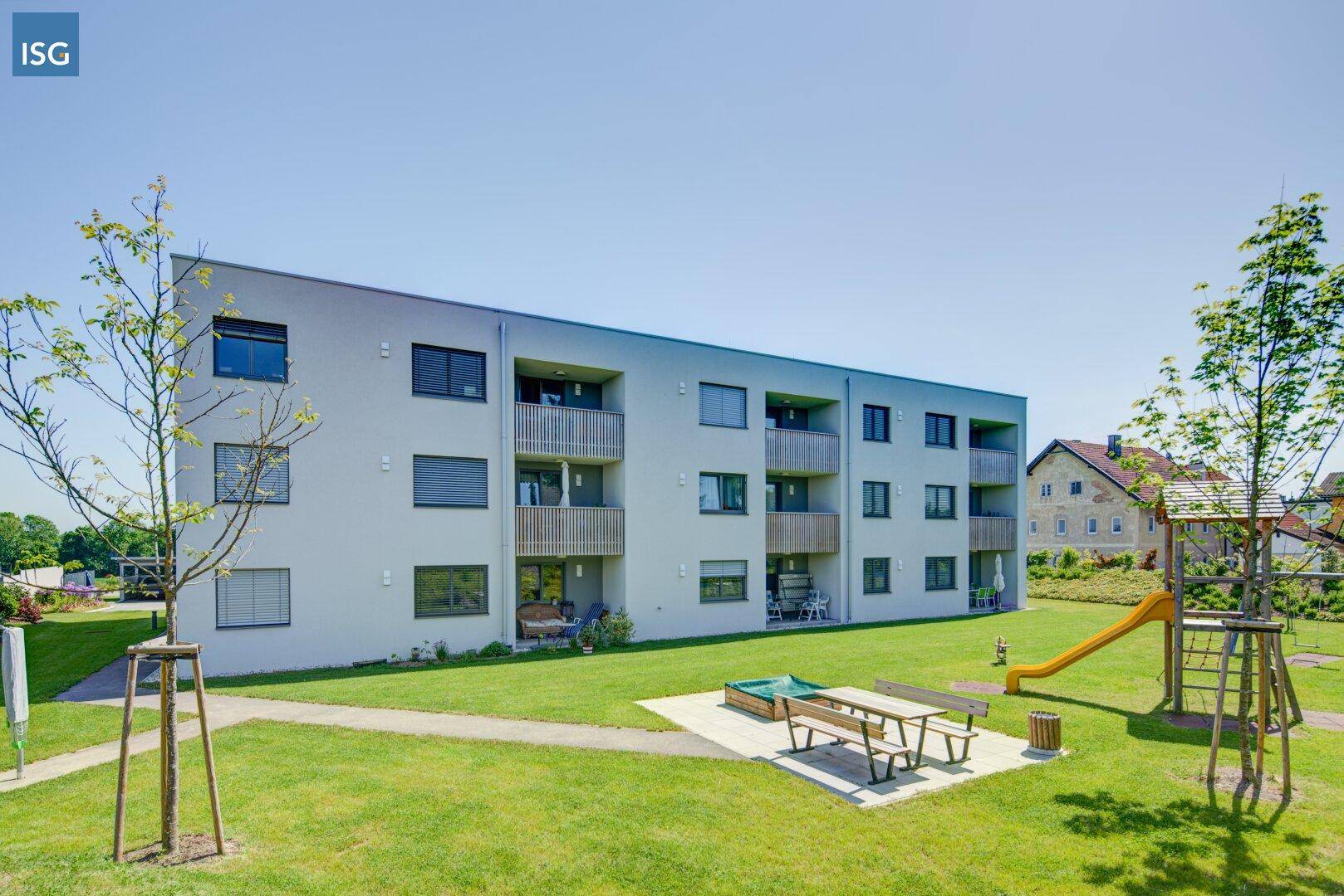 Ansicht eines modernen Wohngebäudes mit Balkonen und einem Gemeinschaftsgarten mit Spielgeräten.