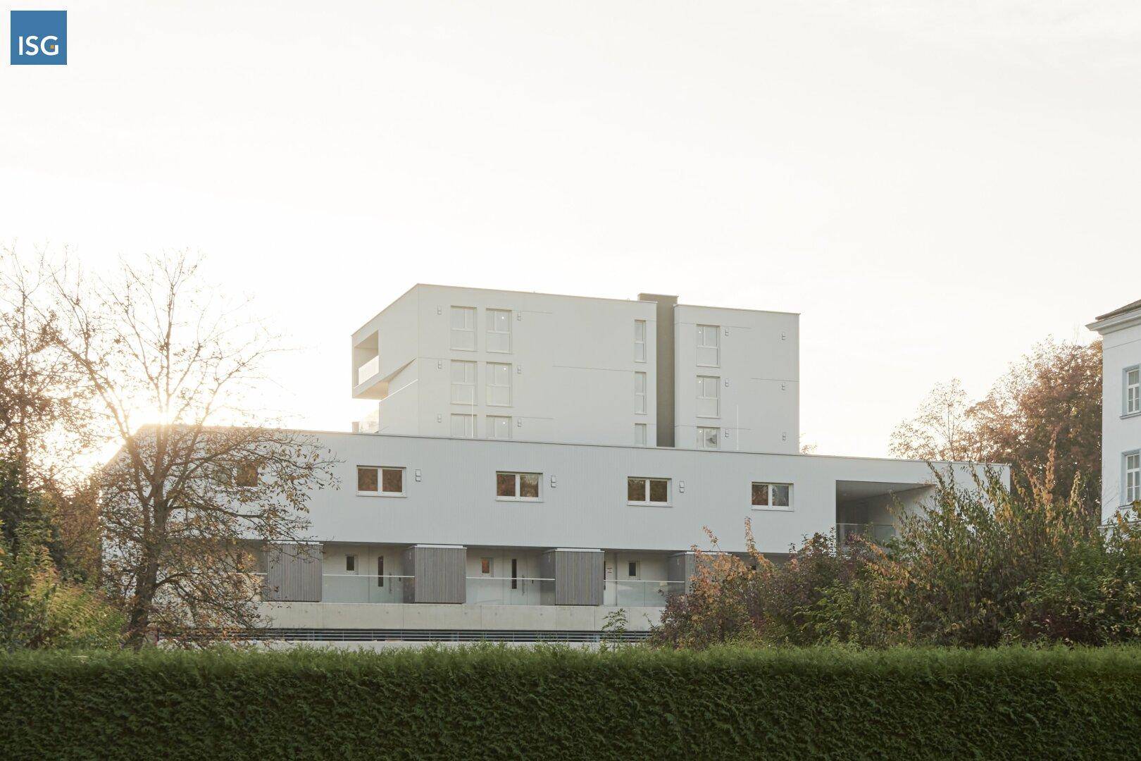Moderne Wohngebäude-Fassade umgeben von Bäumen und einer Hecke im warmen Sonnenlicht.