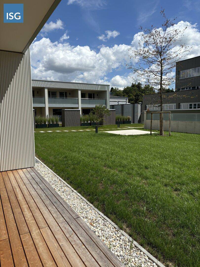 Holzterrasse mit Kiesstreifen und Blick auf eine grüne Rasenfläche und weitere moderne Gebäude.