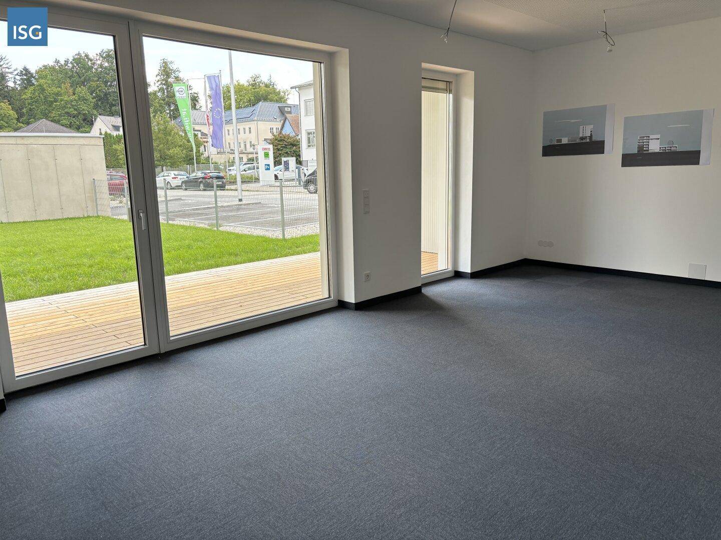Heller Raum mit Teppichboden und großen Fenstern, die auf eine Holzterrasse und die Straße führen.