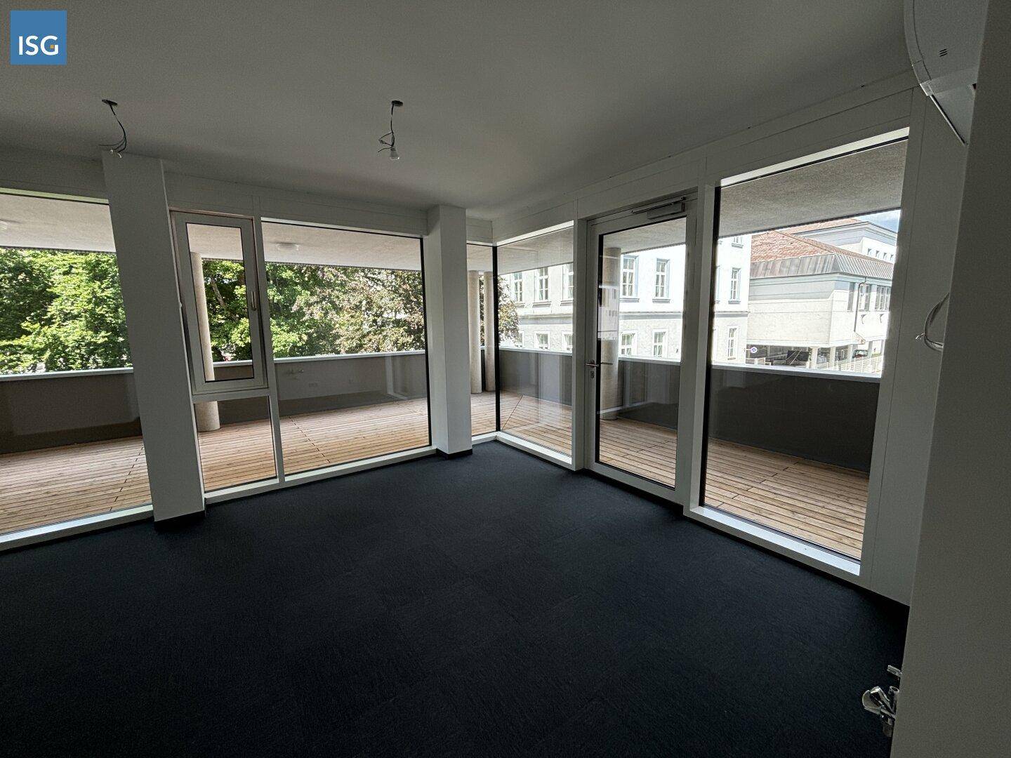 Geräumiger, leerer Raum mit Teppichboden und großen Fensterfronten, die zu einem Balkon führen.
