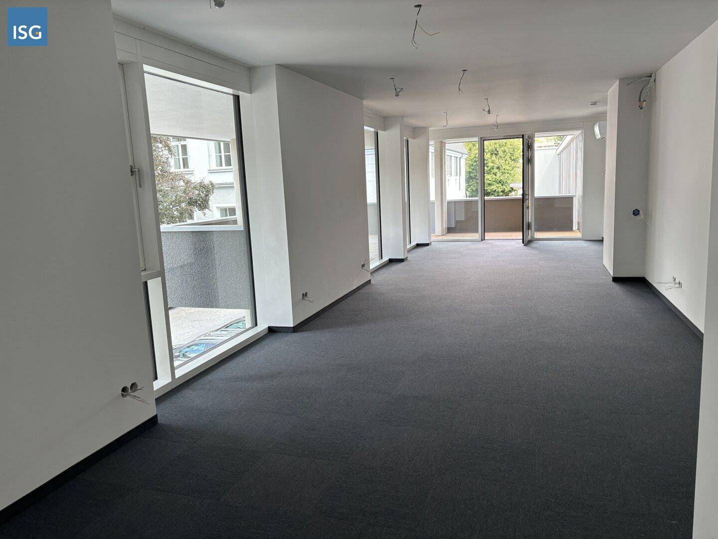 Großer, heller Raum mit Teppichboden und umlaufenden Fensterfronten, die Zugang zu Außenbereichen bieten.