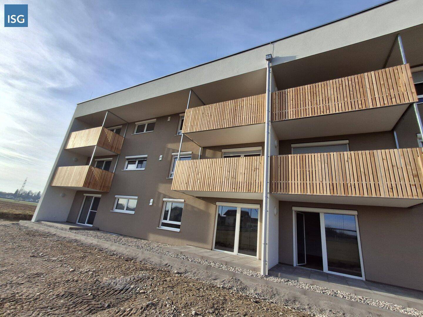 Neubau-Fassade mit mehreren Balkonen und Holzverkleidung, umgeben von unbebautem Gelände.