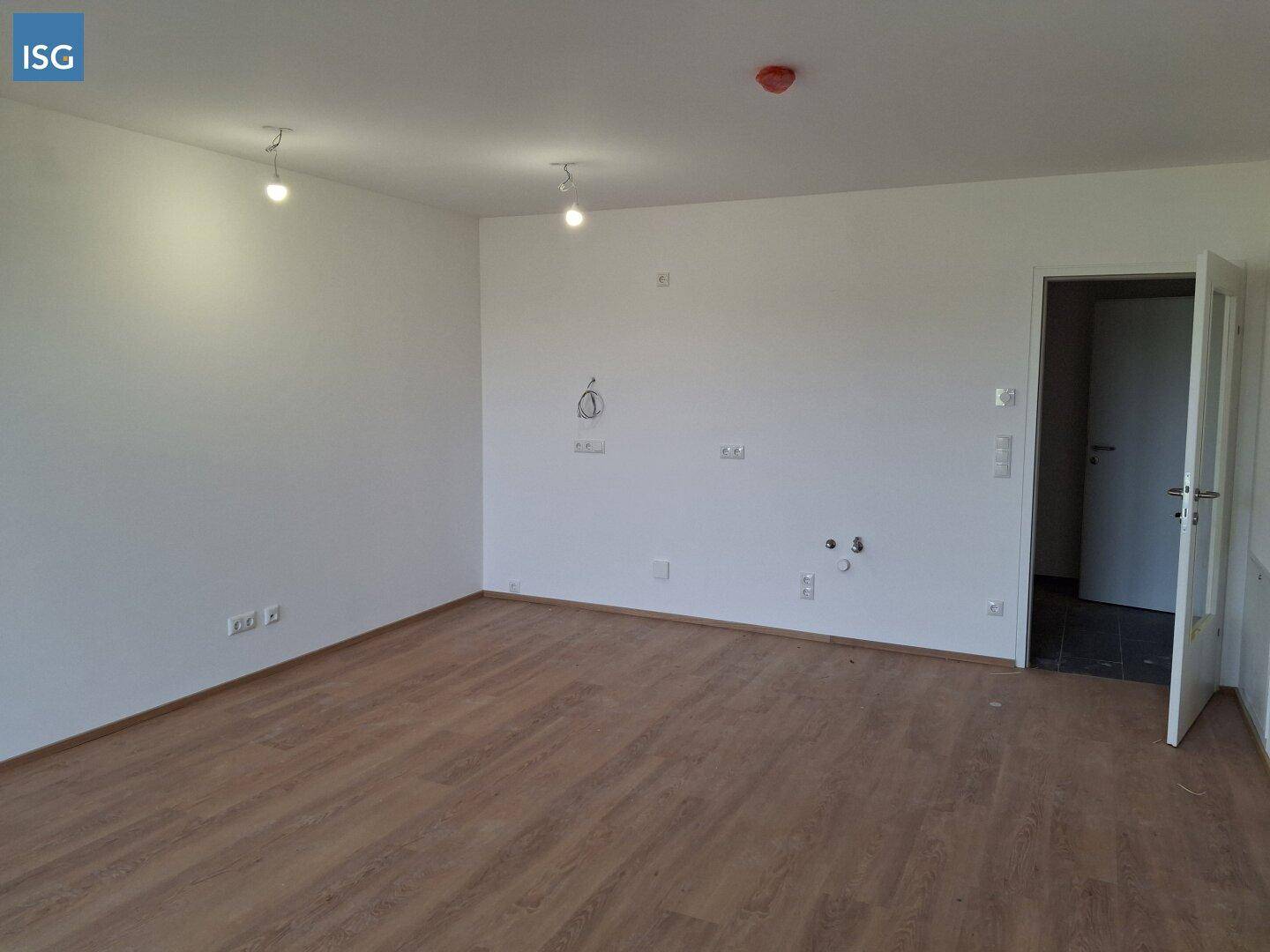 Geräumiger, unmöblierter Raum mit Laminatboden und Anschlüssen für eine zukünftige Küche.