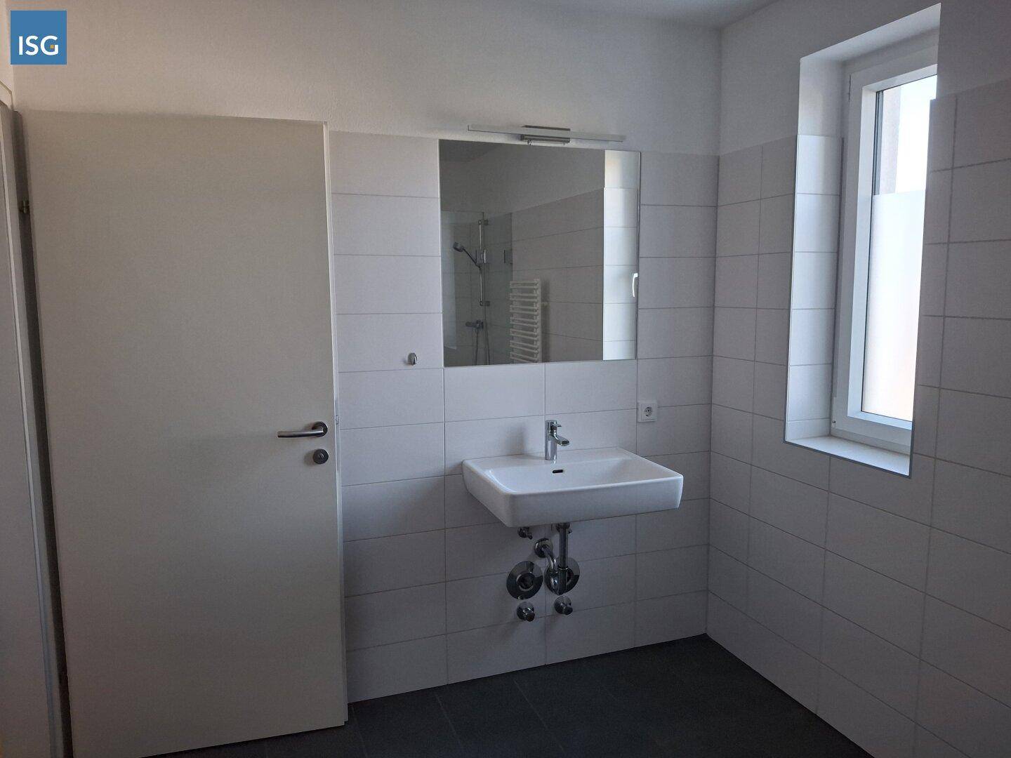 Helles Badezimmer mit Waschbecken, Spiegel und Fenster, ausgestattet mit weißen Fliesen.