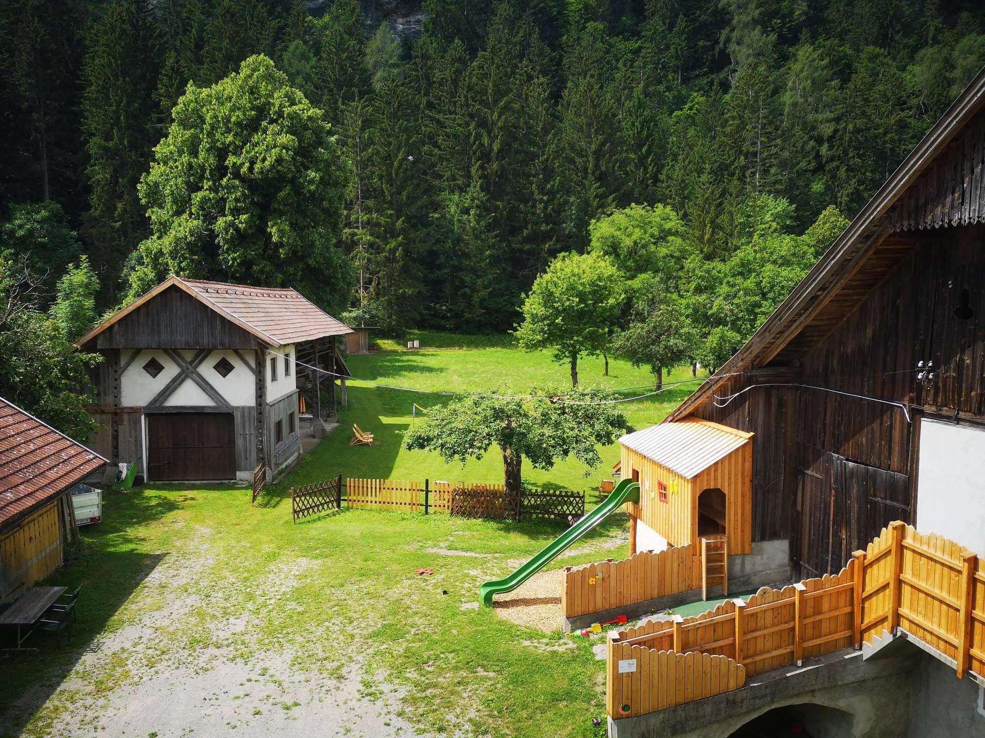 Weitläufiger Außenbereich mit Spielplatz, Rutsche und traditionellen Gebäuden, umgeben von Wald und Natur.