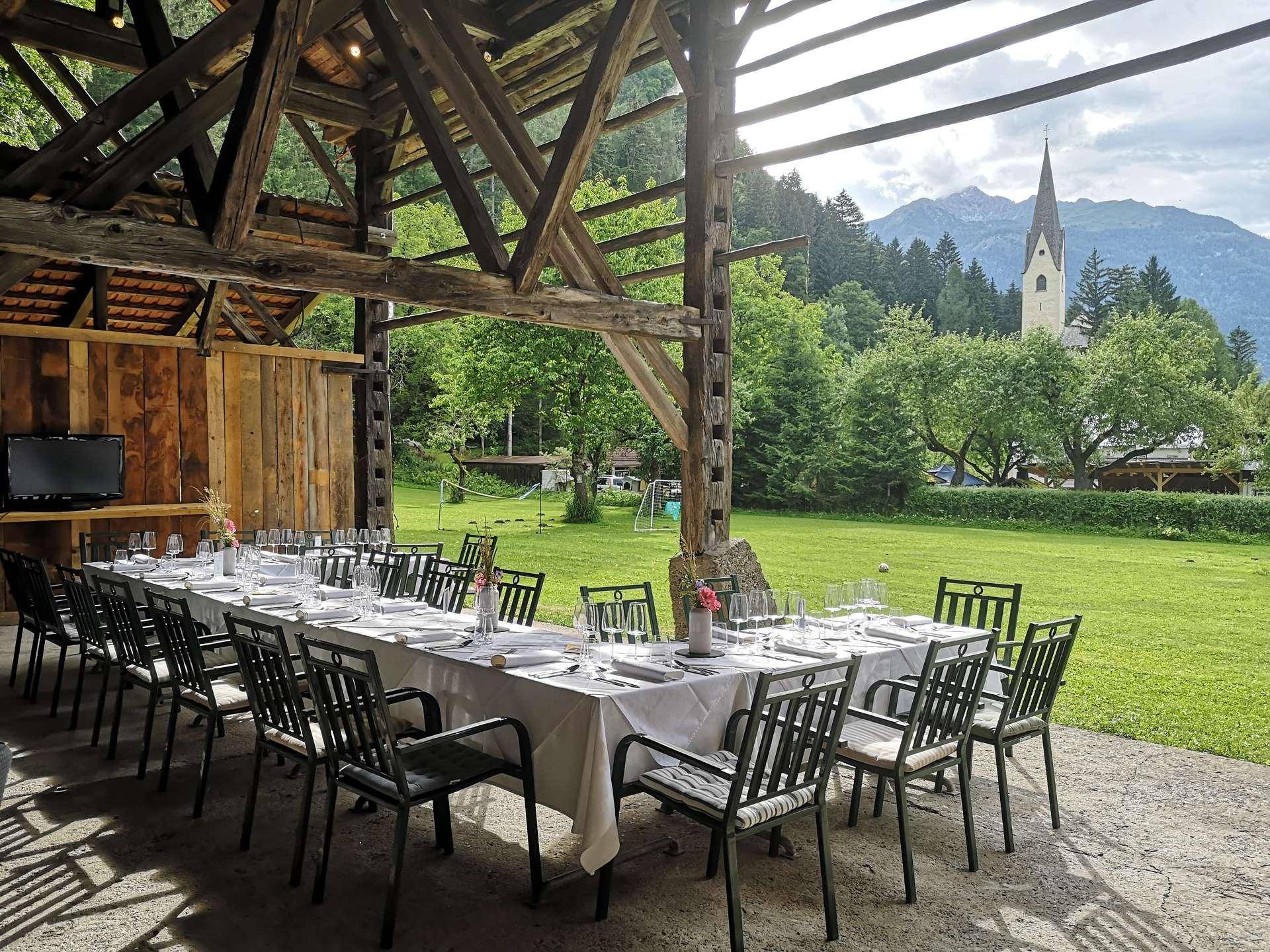 Festlich gedeckter Tisch in einem offenen Holzbau mit Blick auf eine Kirche und die umliegenden Berge.
