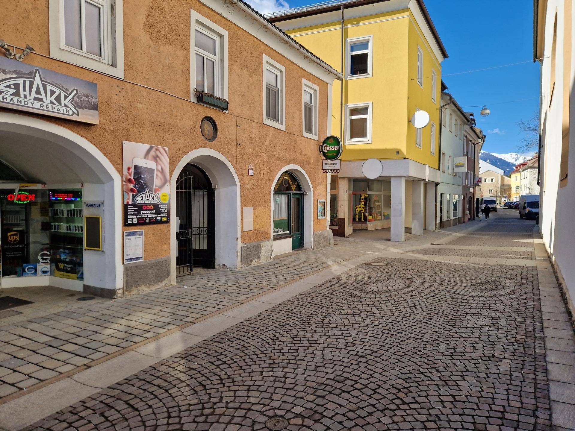 Belebte Kopfsteinpflasterstraße mit historischen Gebäuden und Blick auf schneebedeckte Berge im Hintergrund an einem sonnigen Tag.
