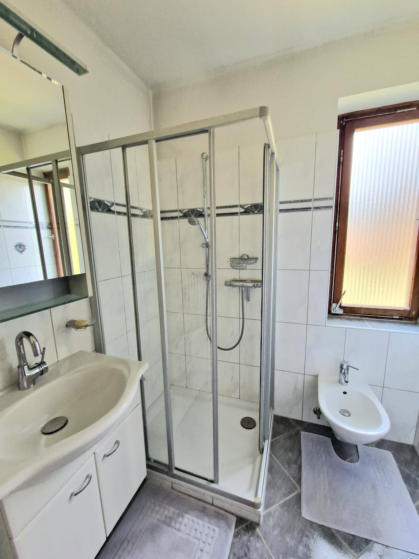 Helles Badezimmer mit Duschkabine, Waschbecken, Bidet und Fenster für natürliche Belüftung.
