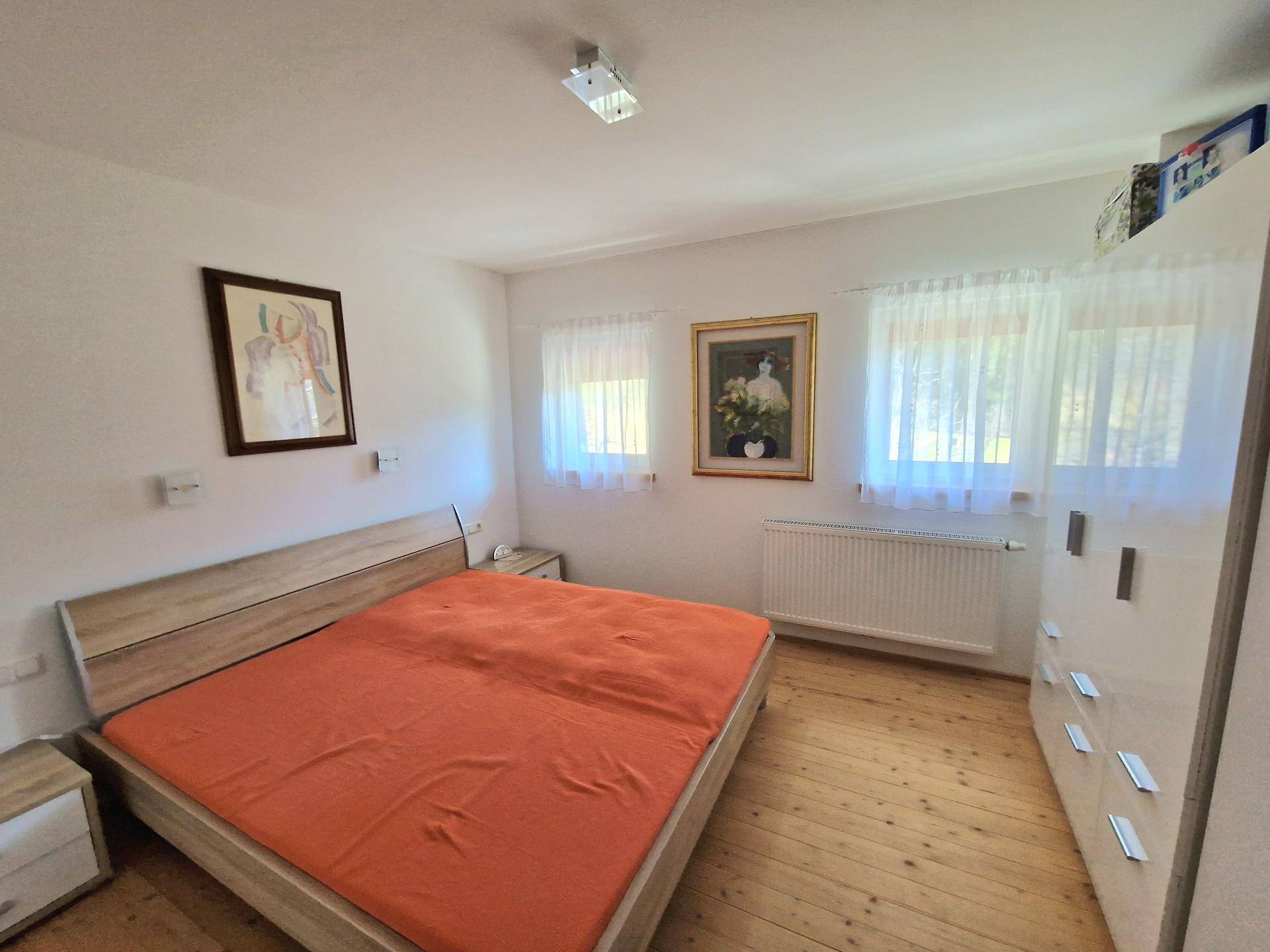 Helles Schlafzimmer mit Holzboden, einem Doppelbett und weißen Vorhängen an den Fenstern.