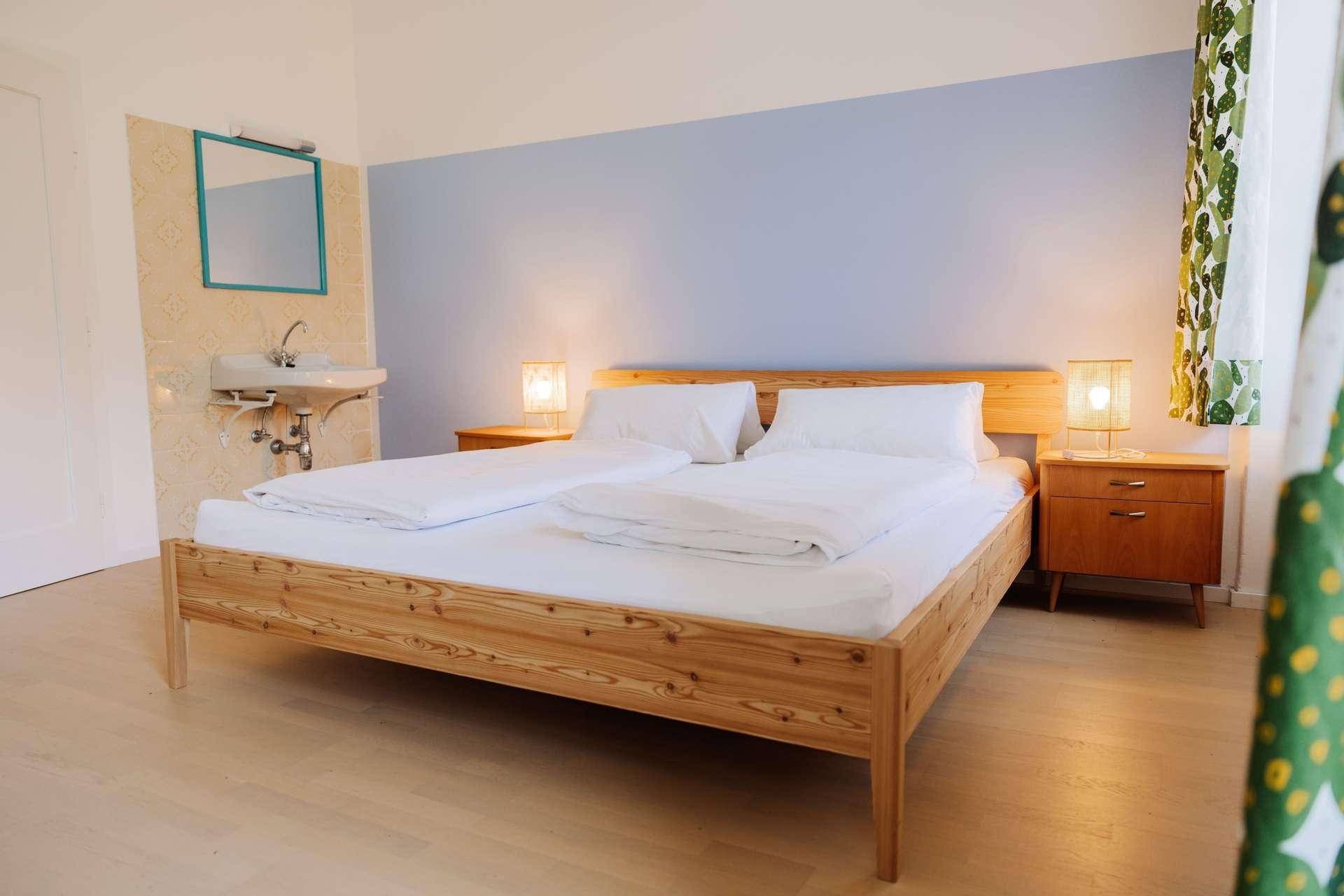 Helles Schlafzimmer mit Holzbett, Nachttischen und einem kleinen Waschbecken in der Ecke des Raumes.