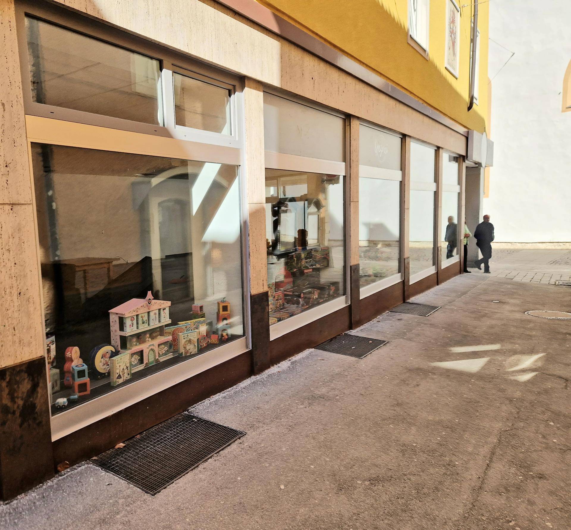 Nahaufnahme der Schaufenster eines Geschäfts mit ausgestellten Waren und Passanten im Hintergrund auf der Gasse.