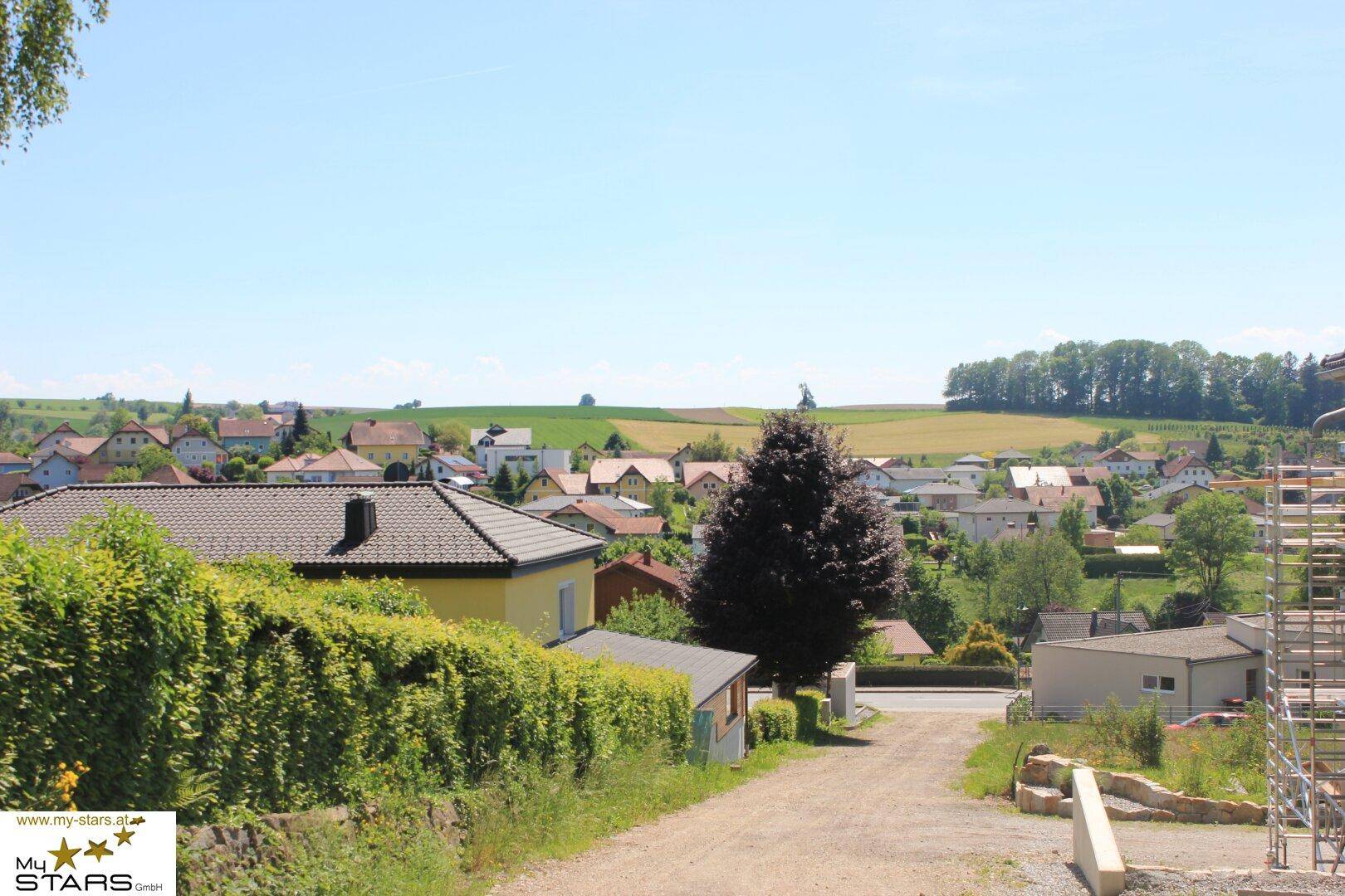 Weitläufiger Blick über ein Dorf mit Feldern und grünen Hügeln unter blauem Himmel.