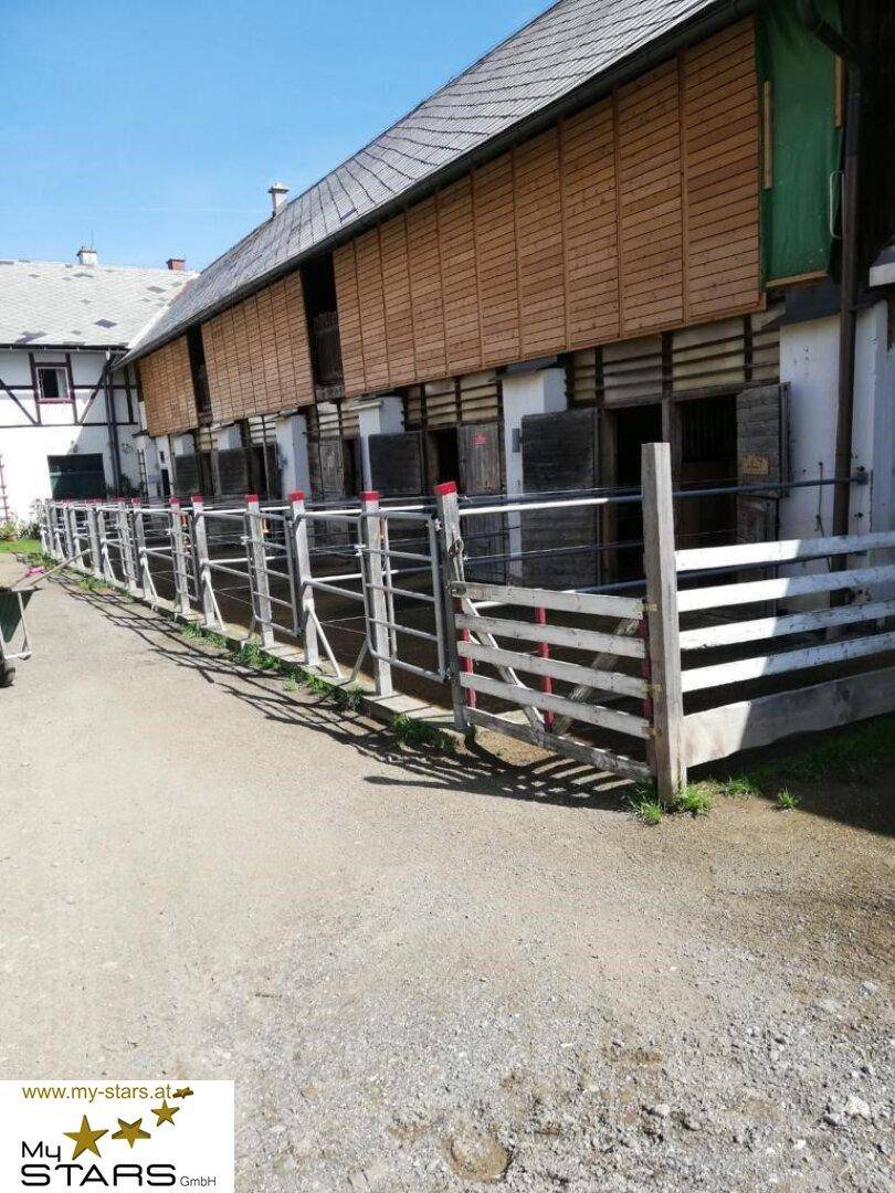 Außenansicht eines Stallgebäudes mit Holzzaun und offenen Boxen in ländlicher Umgebung.
