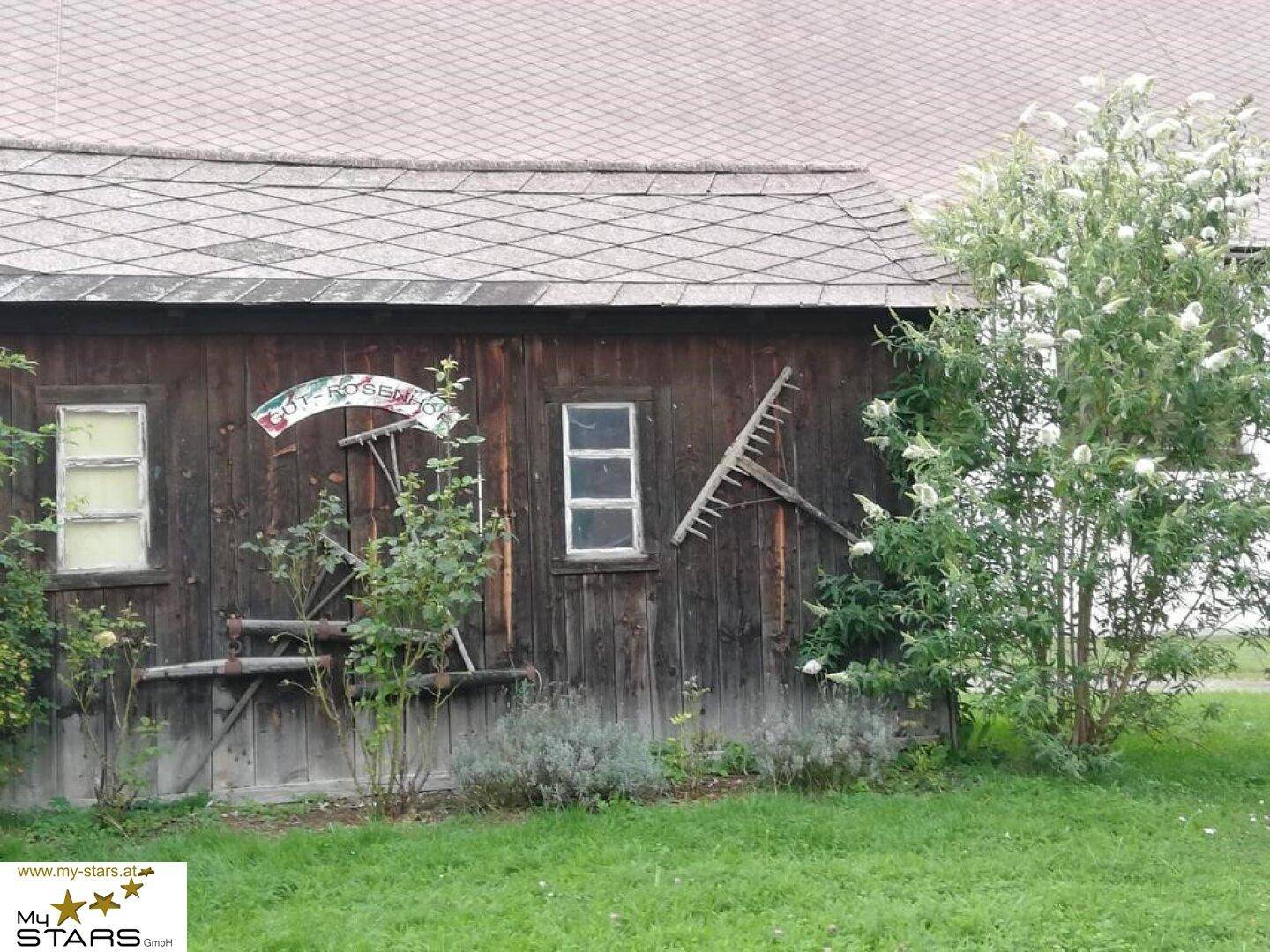 Rustikale Holzwand eines alten Gebäudes mit traditionellen Gartengeräten und blühenden Pflanzen.