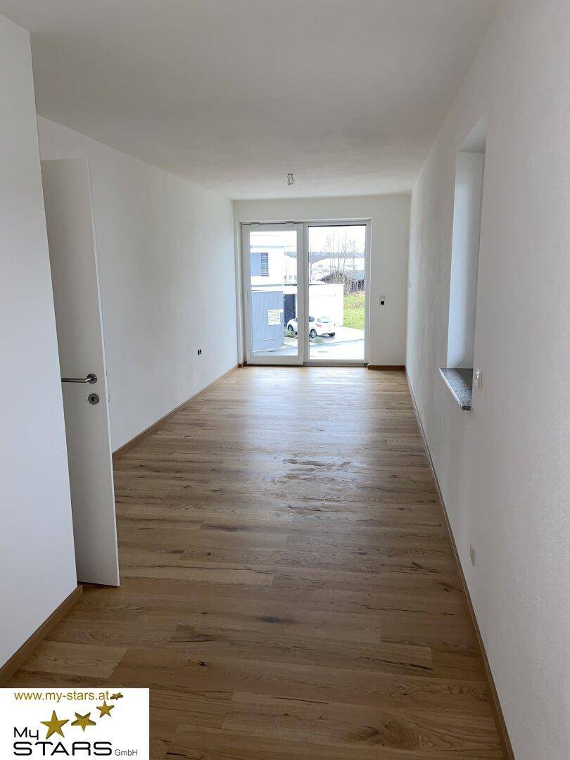 Langer, heller Raum mit Holzboden und einer Glastür, die ins Freie führt.
