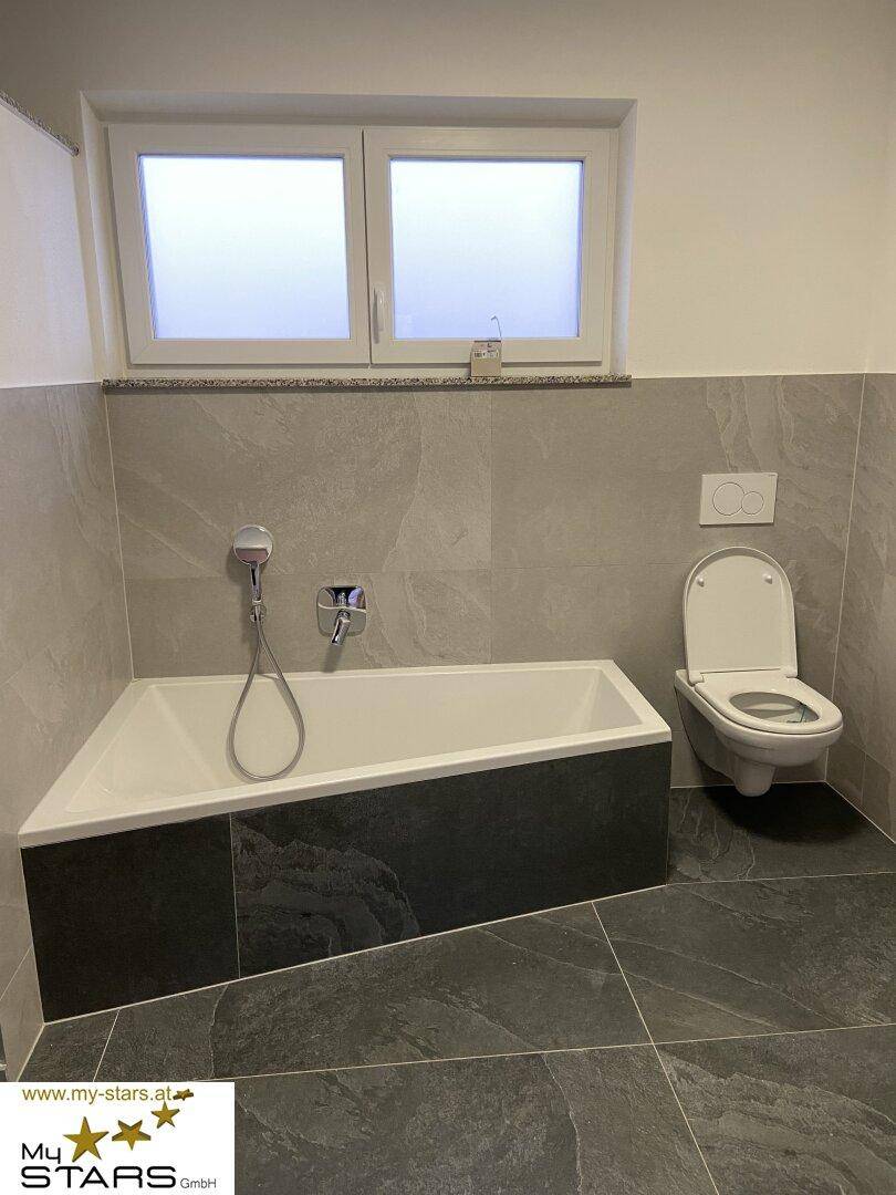 Modernes Badezimmer mit Badewanne, Wand-WC und großformatigen grauen Fliesen sowie einem Fenster.