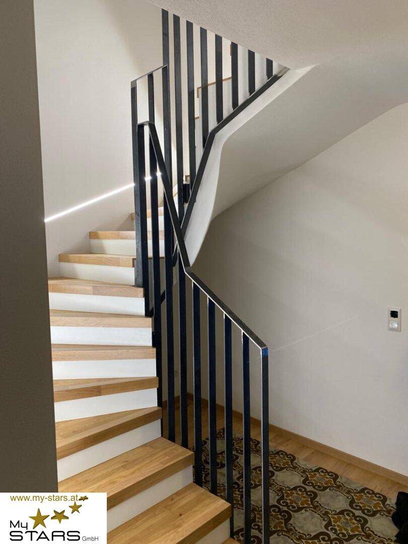Moderne Treppe mit Holzstufen und elegantem schwarzen Metallgeländer, gut beleuchtet.