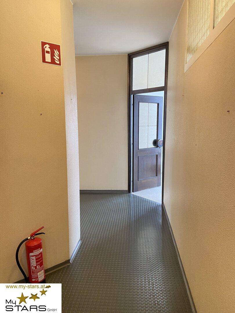 Schmaler Flur mit dunklem Gummiboden, beigen Wänden und einem Feuerlöscher in der Ecke.