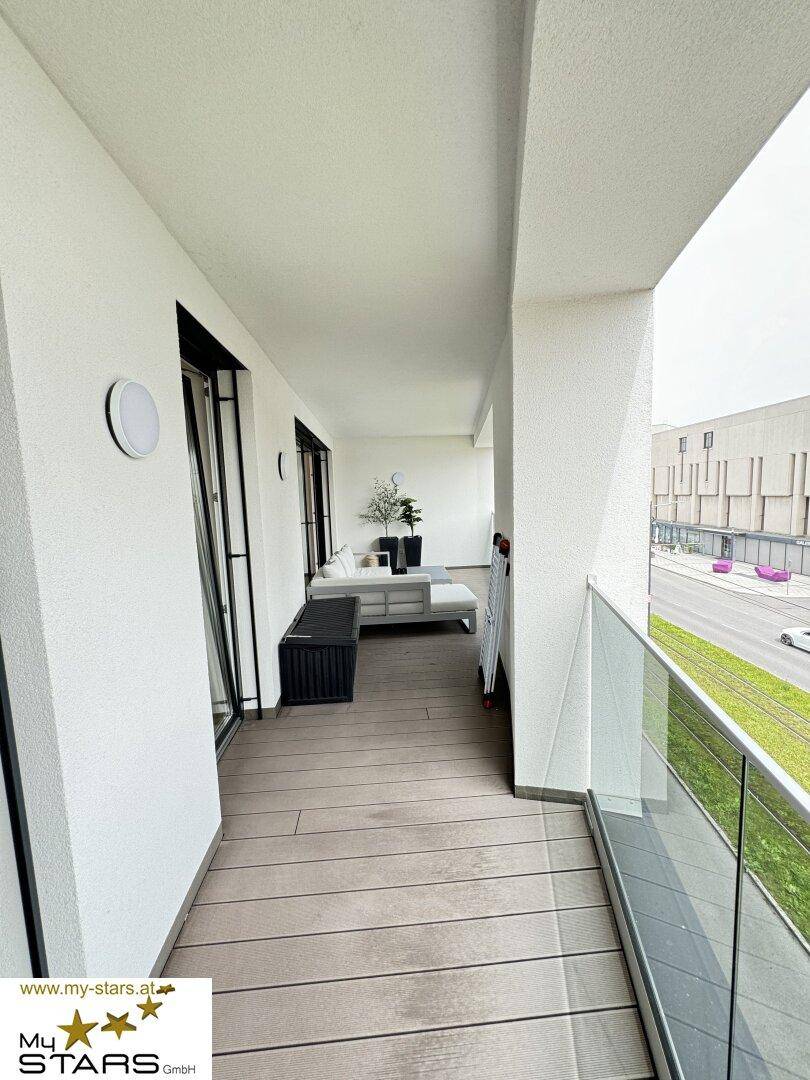 Langer Balkon mit Holzfußboden, Glasgeländer und Blick auf die Straße.
