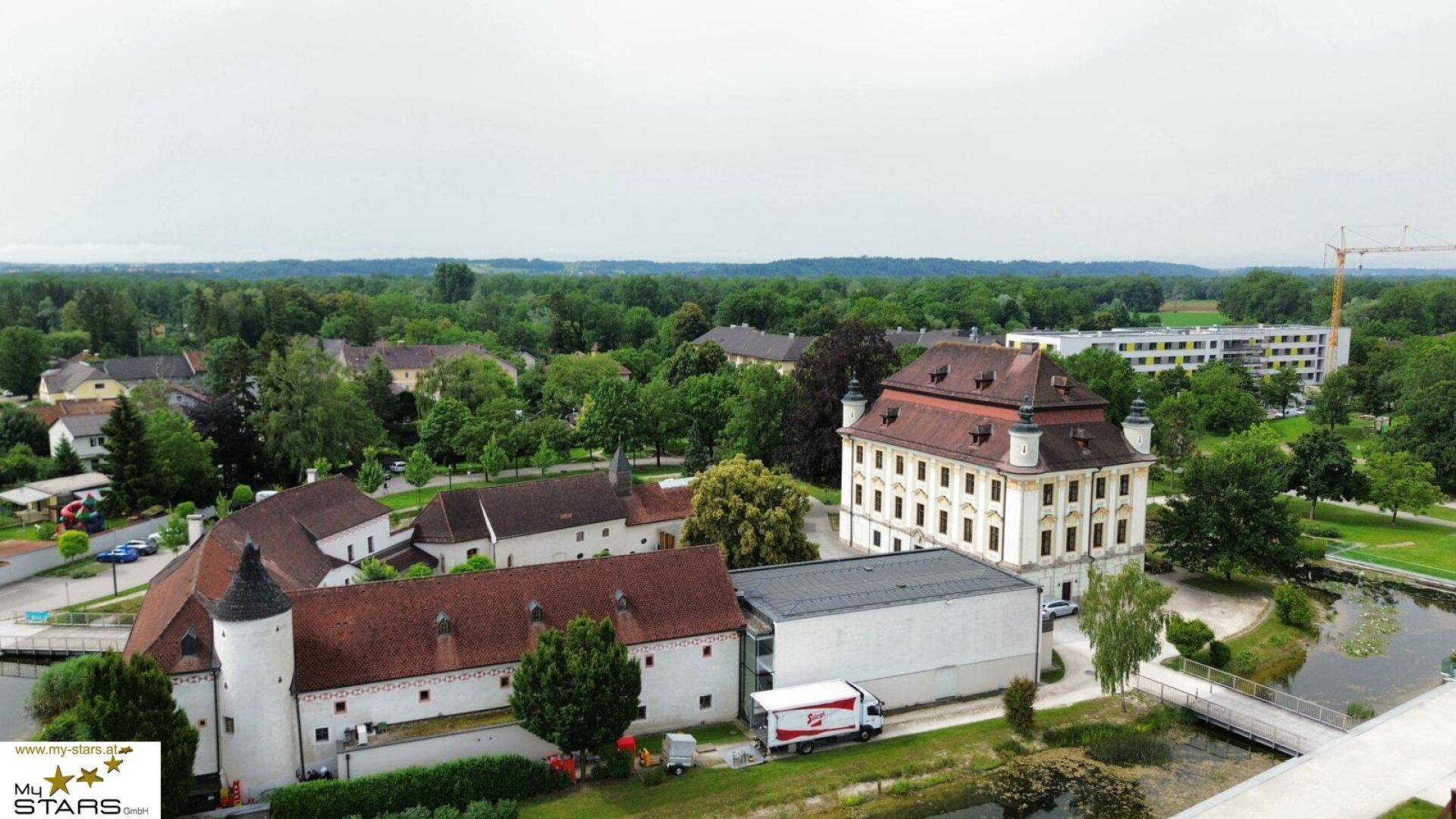 Luftaufnahme eines historischen Gebäudes mit Parkanlage und Wasserlauf in ländlicher Umgebung.