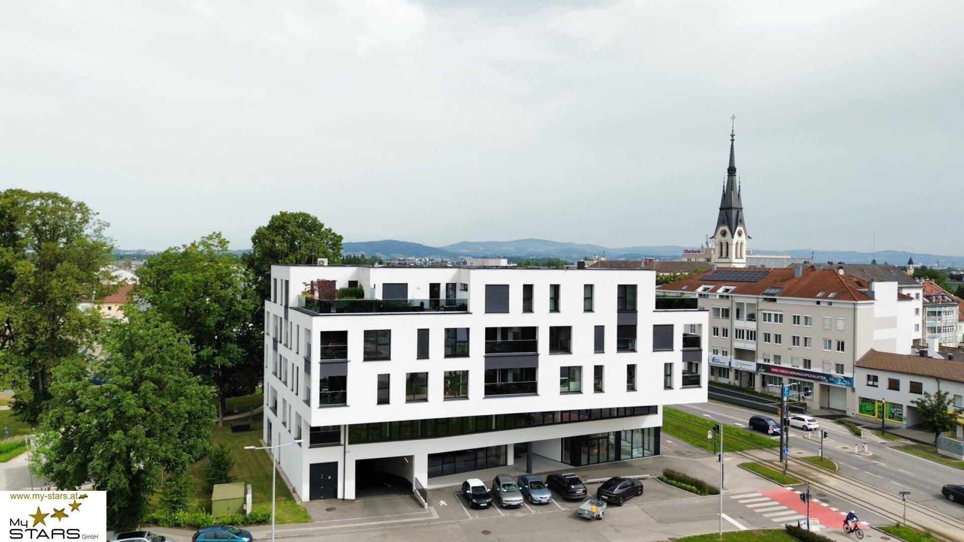 Luftaufnahme eines modernen Gebäudes in städtischer Umgebung mit Kirchturm im Hintergrund.