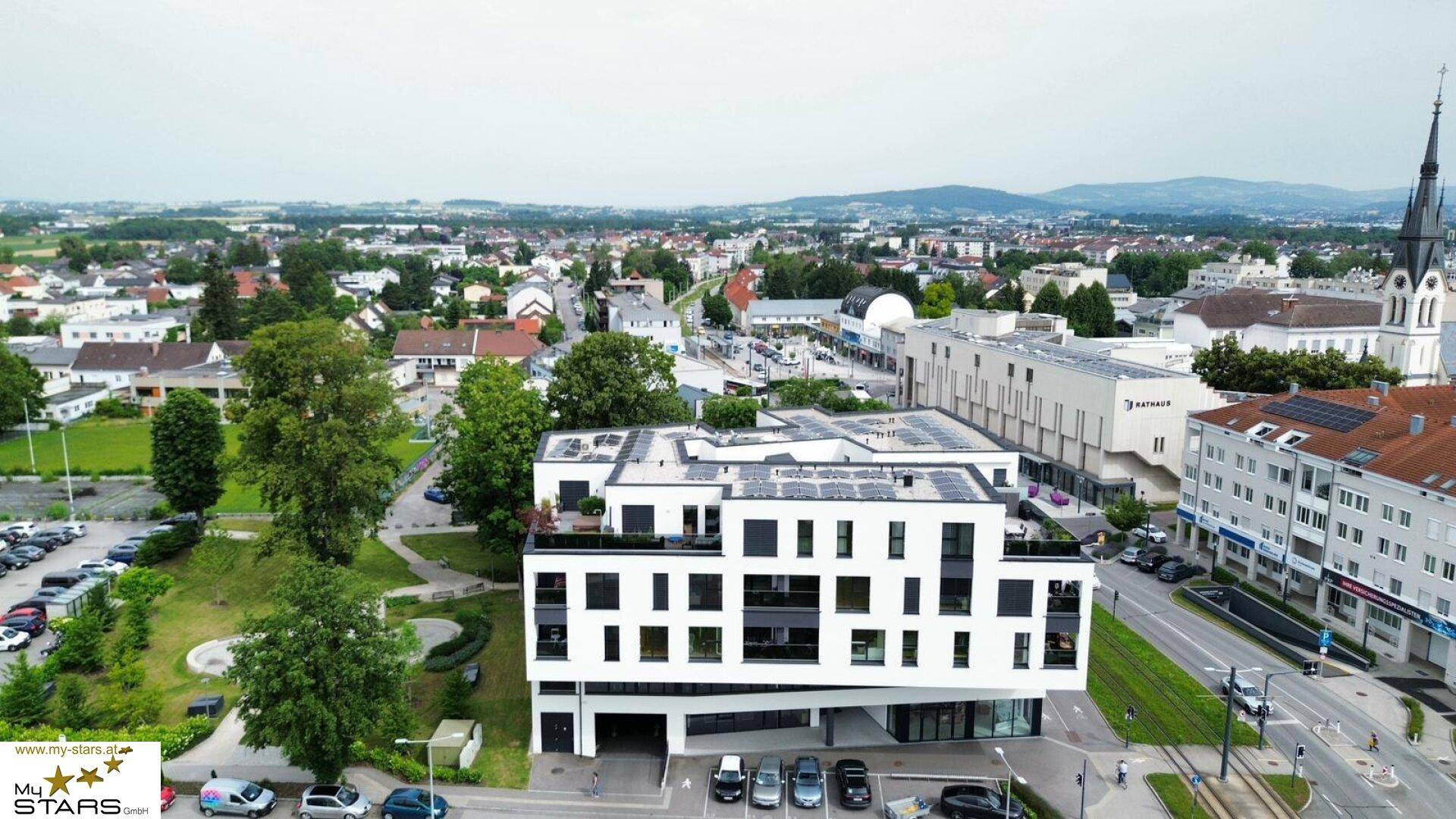 Luftaufnahme eines modernen Gebäudes mit Solarpaneelen auf dem Dach und städtischer Umgebung.