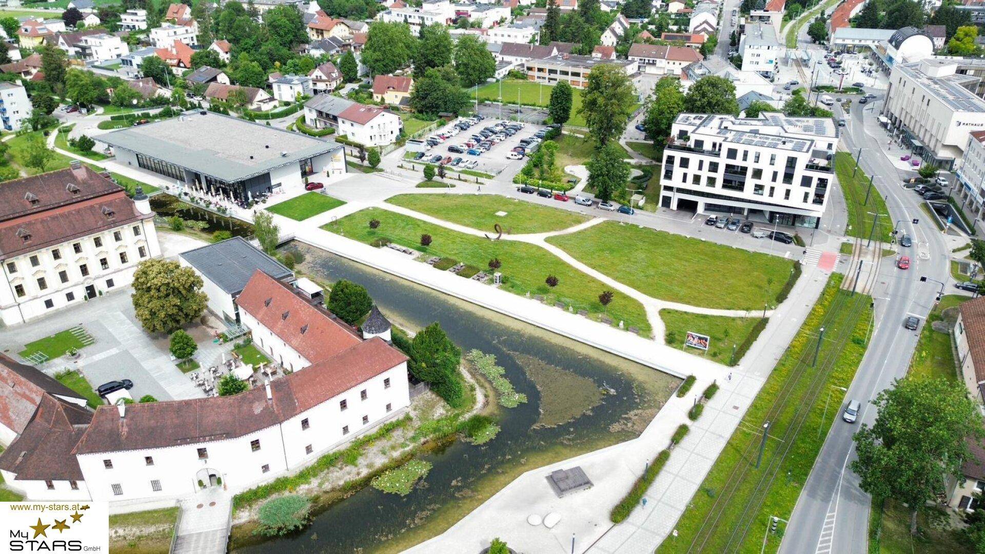 Luftaufnahme eines modernen Gebäudes und einer historischen Anlage mit Grünflächen und Wasser.