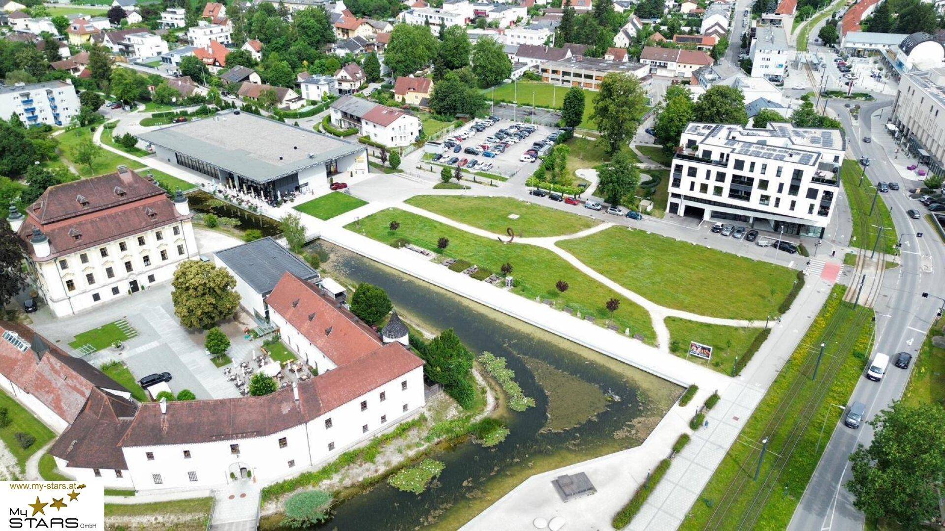 Historische Gebäude entlang eines Kanals, umgeben von weitläufigen Grünflächen und einer modernen Wohnanlage im Hintergrund.