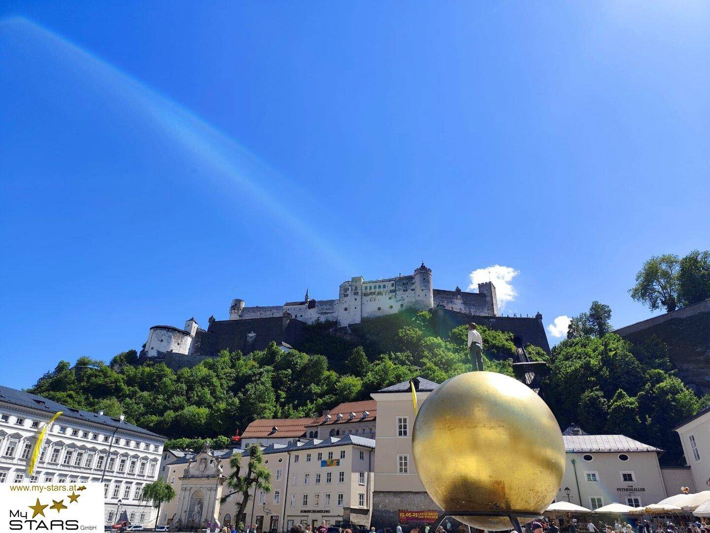 Historischer Stadtplatz in Salzburg mit der Festung Hohensalzburg unter blauem Himmel.