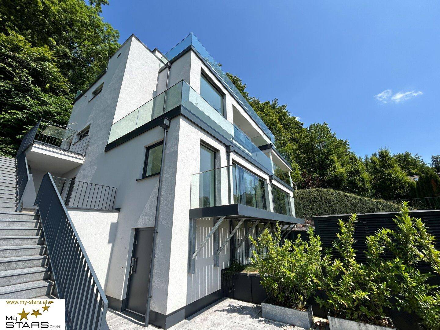 Moderne Architektur mit klaren Linien, Glasbalkonen und Außentreppe in grüner Umgebung.