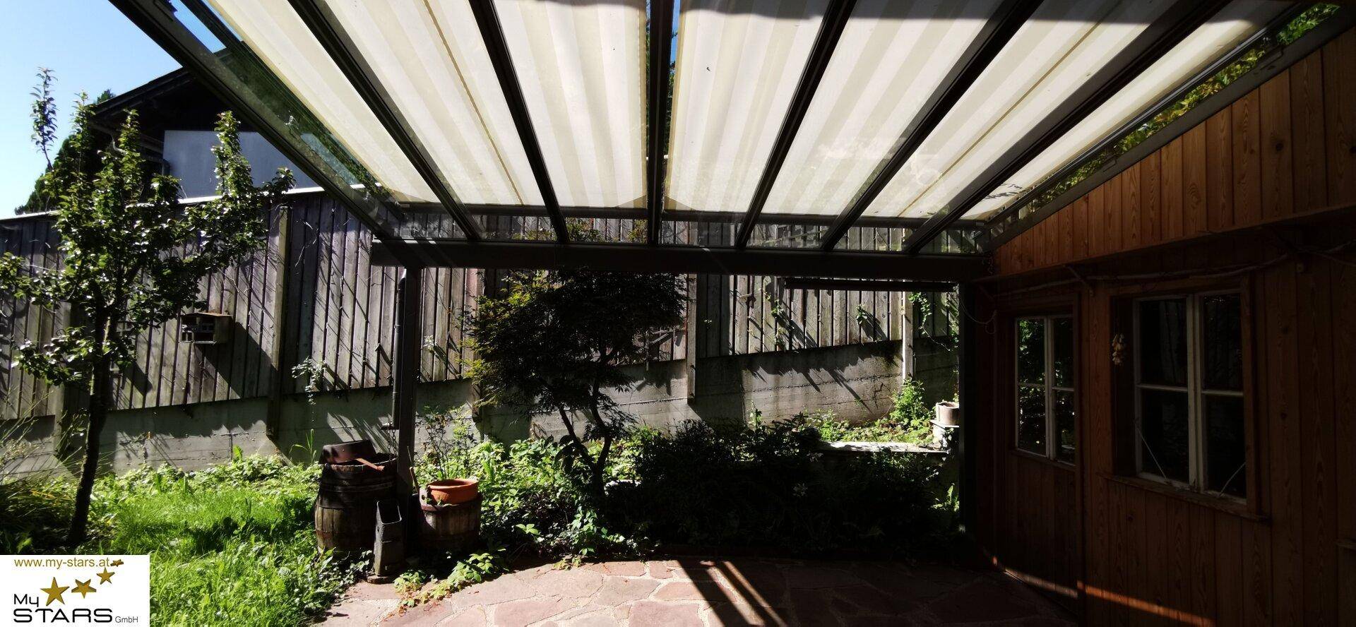 Gartenbereich mit Steinplattenboden unter einer überdachten Pergola, umgeben von Bäumen.