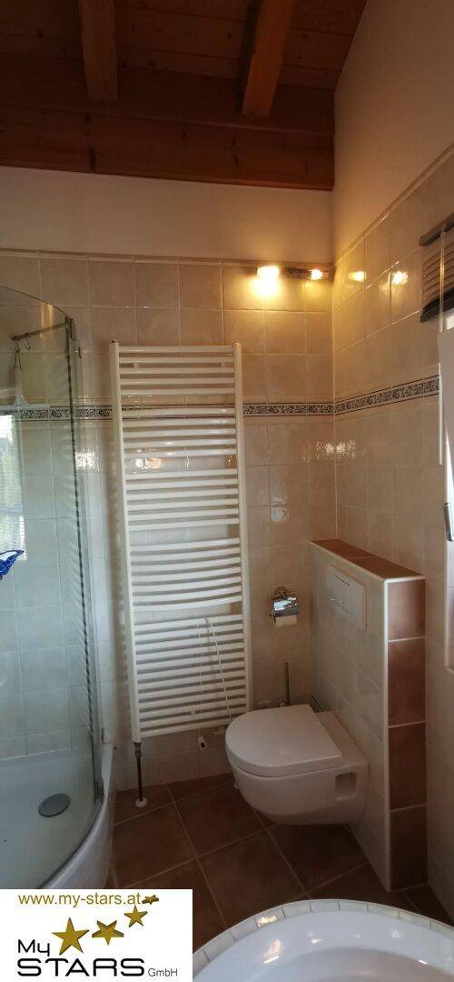 Badezimmer mit Dusche, WC und beheiztem Handtuchhalter, unter einer Holzbalkendecke.