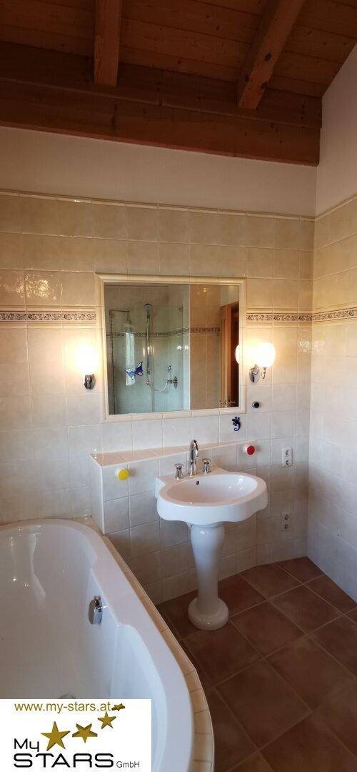 Badezimmer mit Badewanne, Waschbecken und Spiegel, unter einer rustikalen Holzbalkendecke.